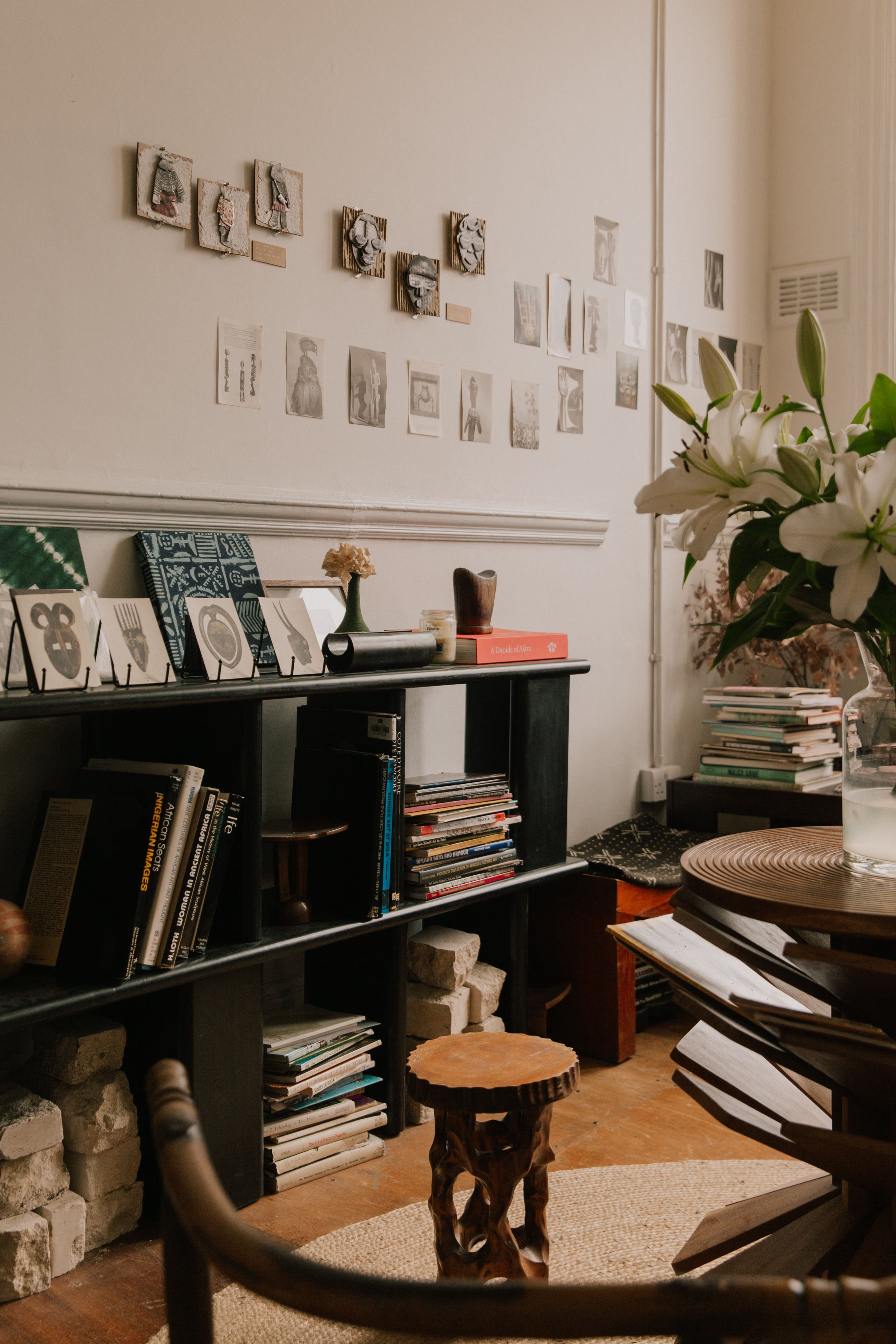 A cozy room features a wall decorated with small artworks and photos, a dark bookshelf filled with books, and white lilies in a vase next to a carved wooden stool.