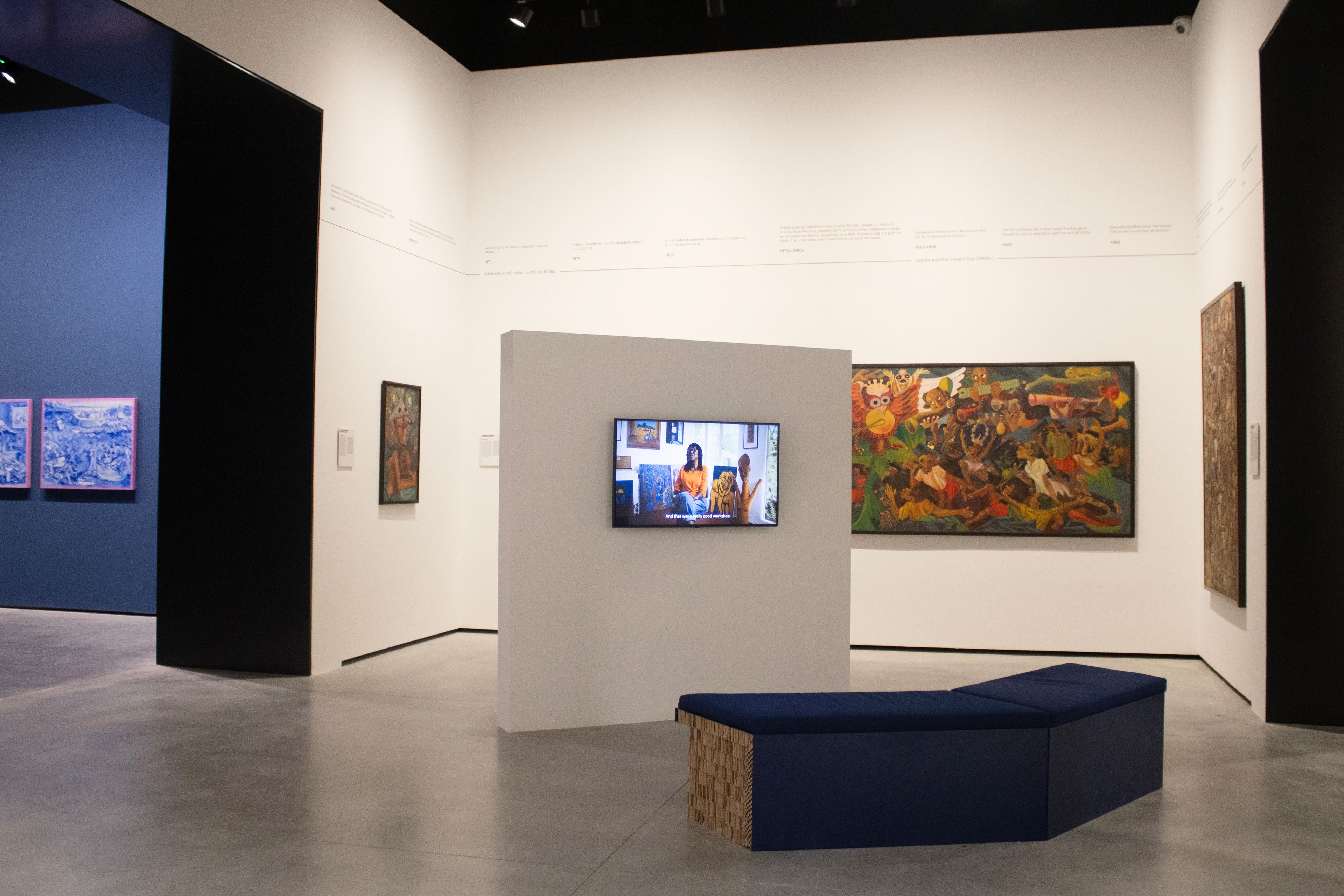 A brightly lit art gallery with white walls, a concrete floor, a large colorful painting, a monitor showing two people on a partition, and a blue L-shaped bench.