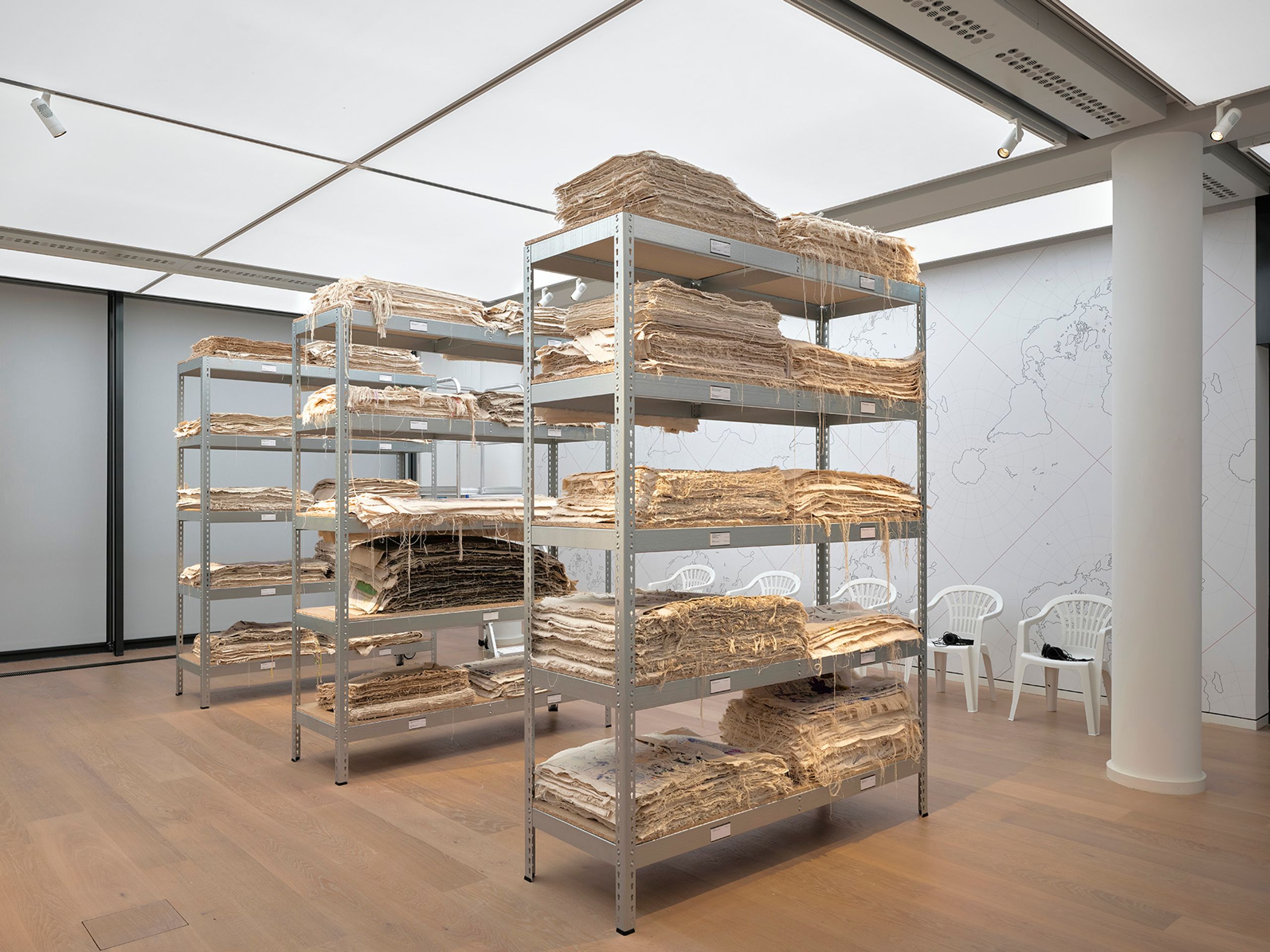 Metal shelves packed with stacks of old, light-colored paper or fabric, in a room with a faint world map on the wall.