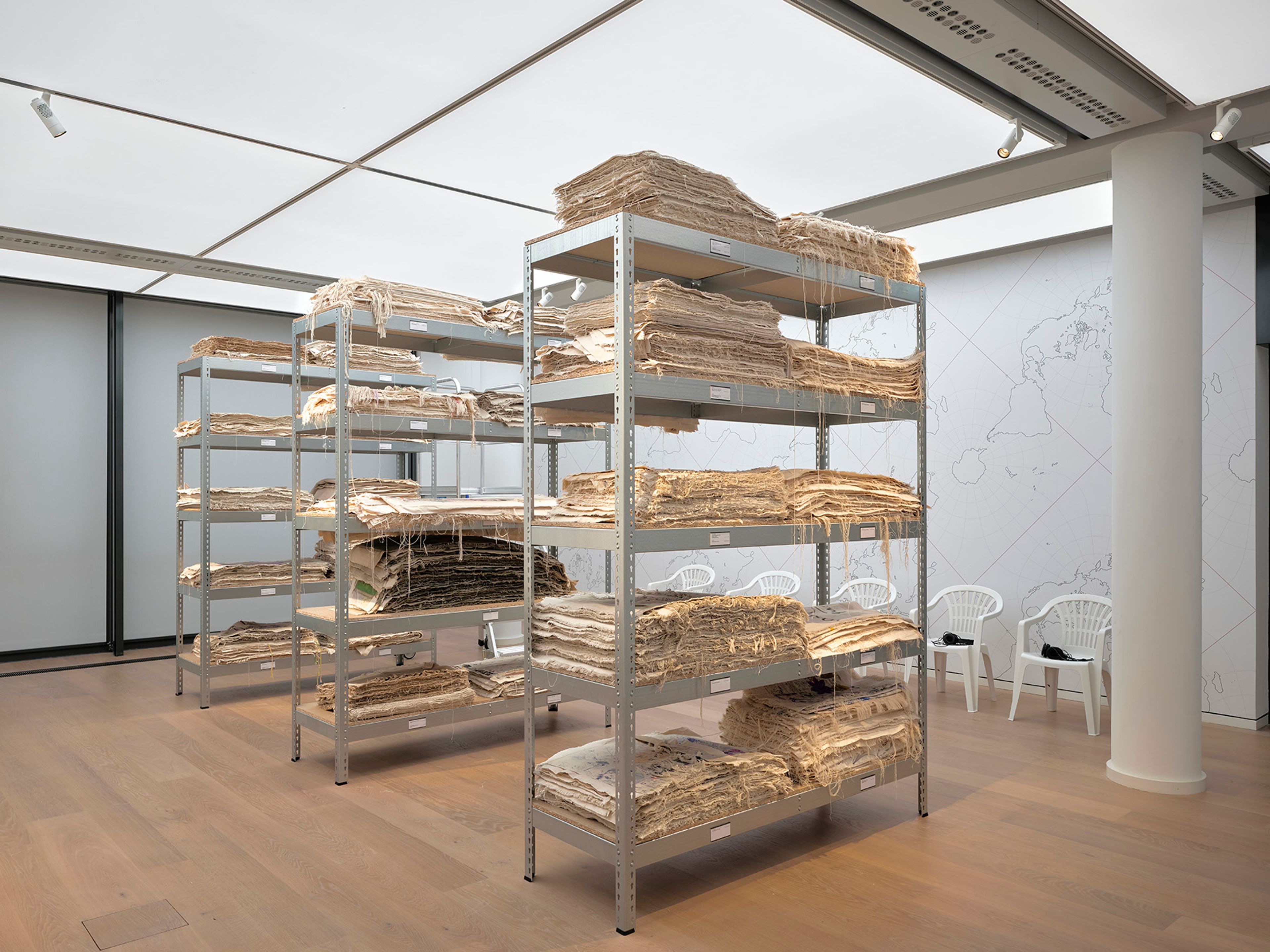 Metal shelves packed with stacks of old, light-colored paper or fabric, in a room with a faint world map on the wall.