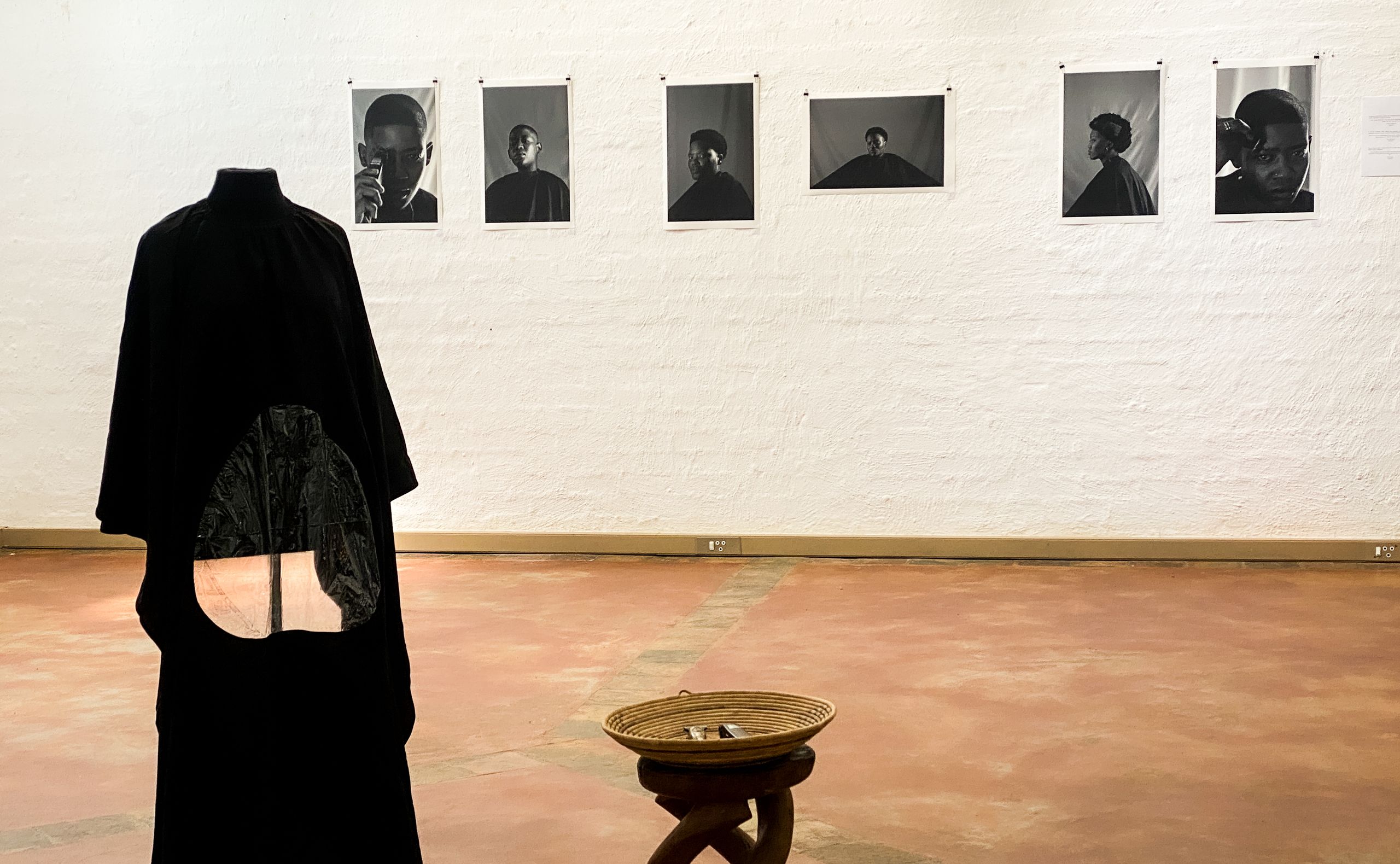 A gallery view featuring a black sculptural garment on a mannequin, a series of black and white portraits on the wall, and a woven basket on a wooden stool.