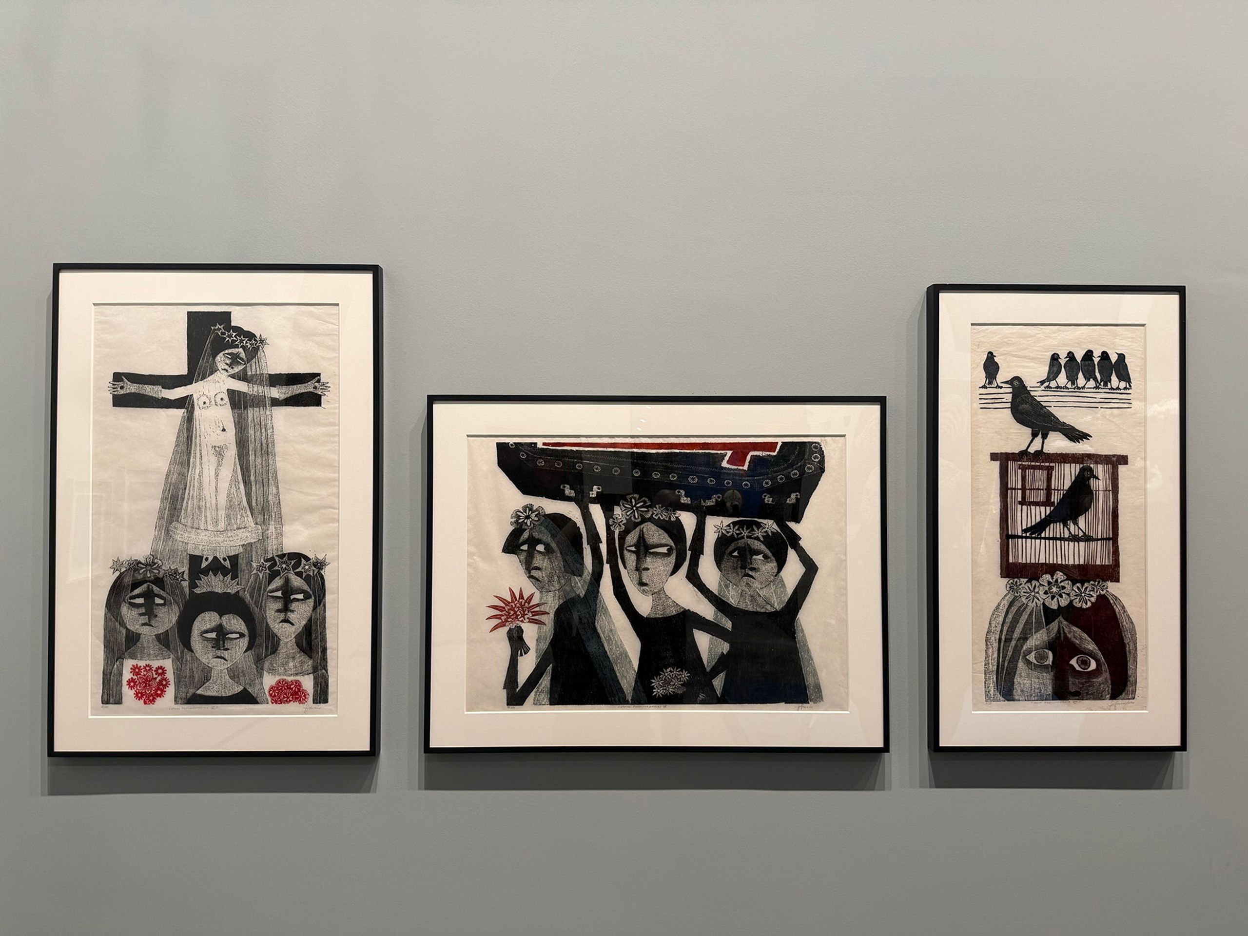 Three framed black, white, and red artworks featuring stylized figures, a crucifixion, and birds, on a grey wall.