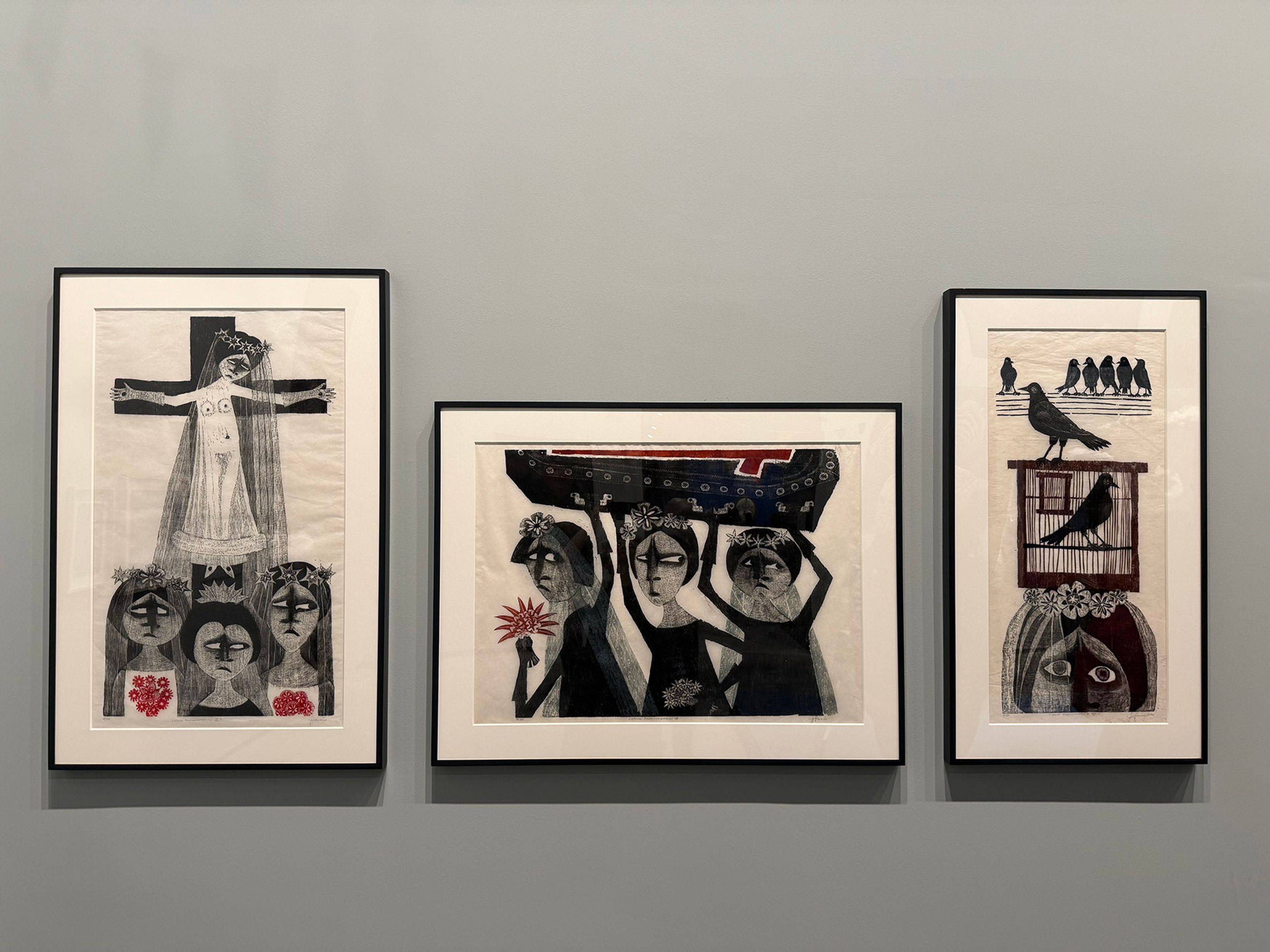 Three framed black, white, and red artworks featuring stylized figures, a crucifixion, and birds, on a grey wall.