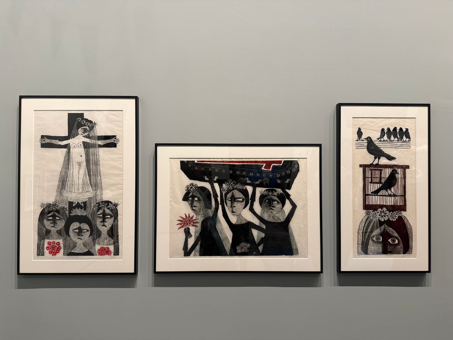Three framed black, white, and red artworks featuring stylized figures, a crucifixion, and birds, on a grey wall.