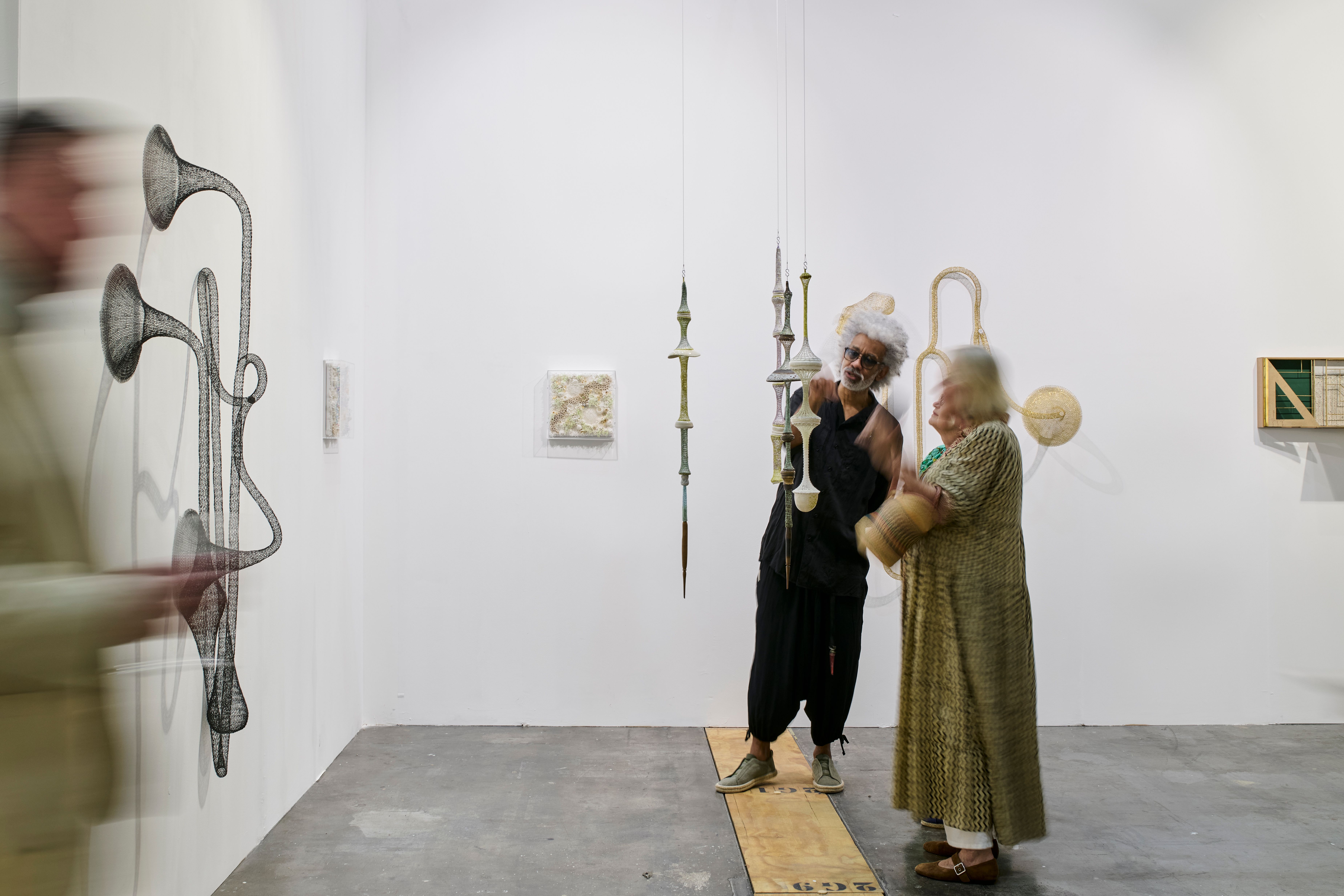 A man adjusts a hanging sculpture in a bright art gallery as a woman watches, with a blurred figure viewing a wall sculpture nearby.