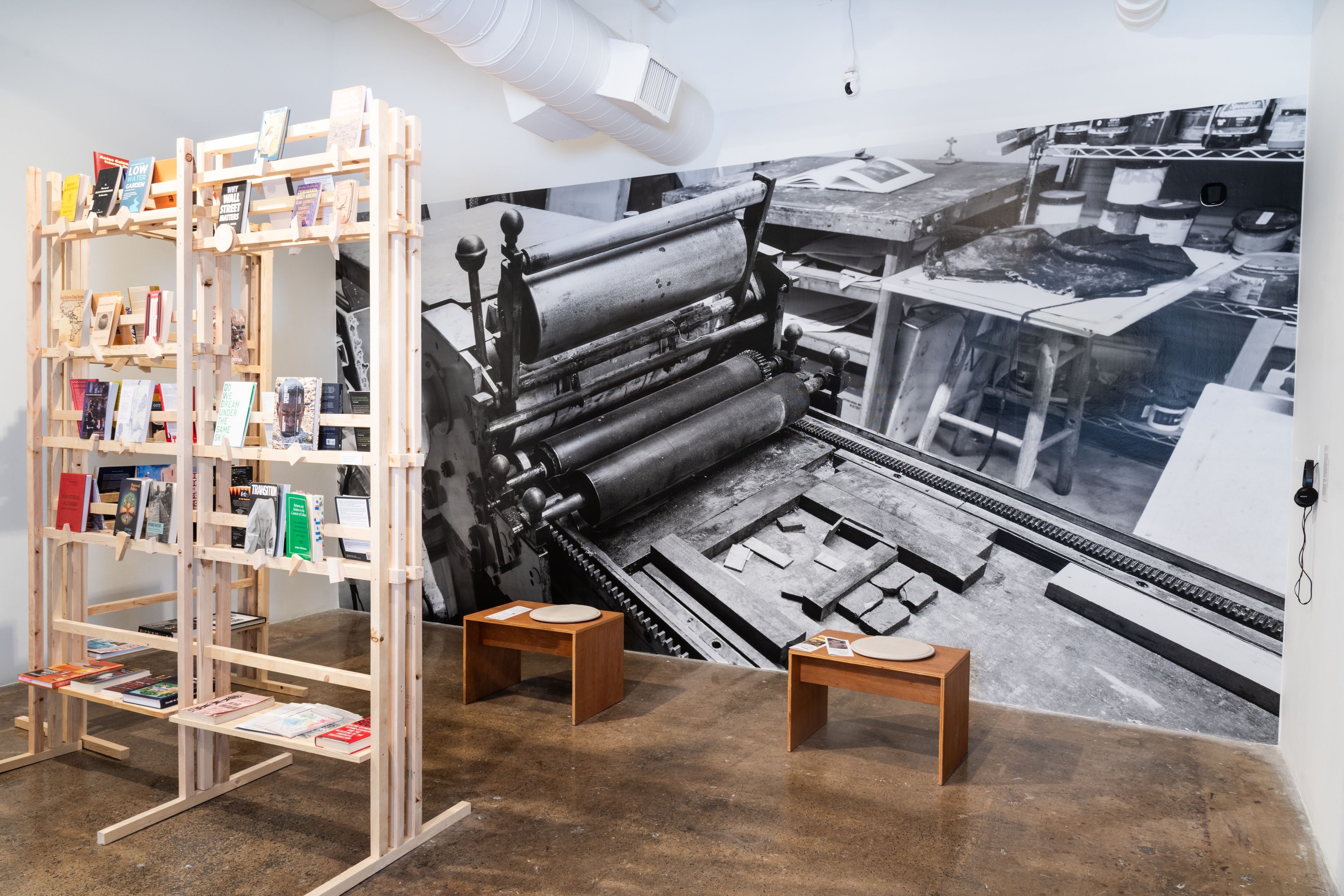 A room with a wooden bookshelf filled with books, two small tables, and a large black and white mural of a vintage printing press and workshop.
