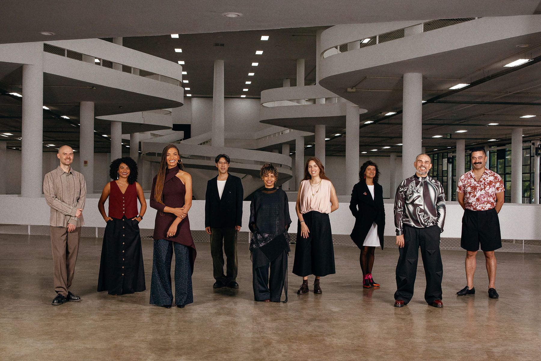 Nine diverse individuals in varied outfits pose in a large, modern architectural building with a curved interior ramp.