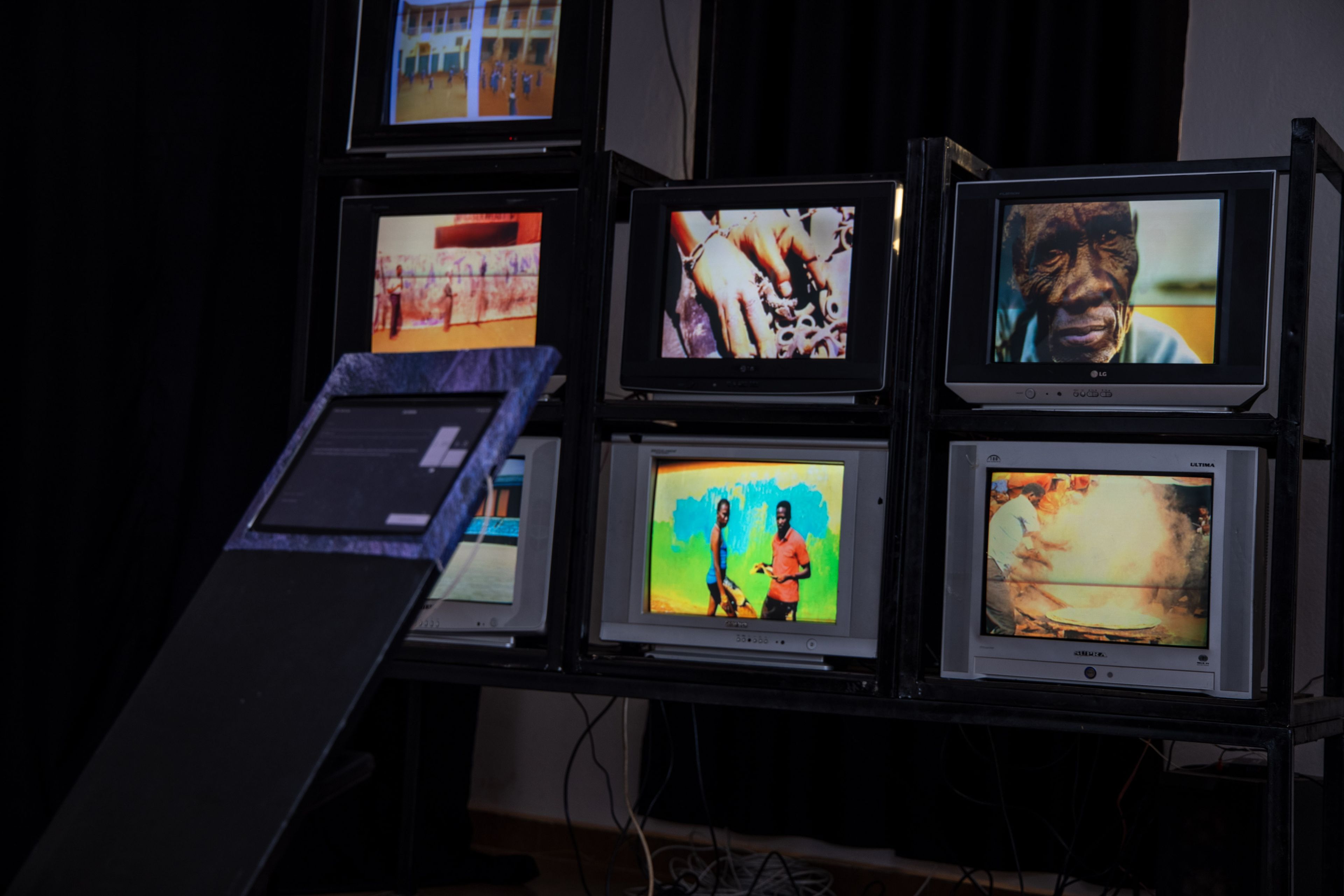 A contemporary art installation featuring seven CRT televisions stacked on shelves, each displaying a different image or video, alongside a tilted interactive screen on the left.