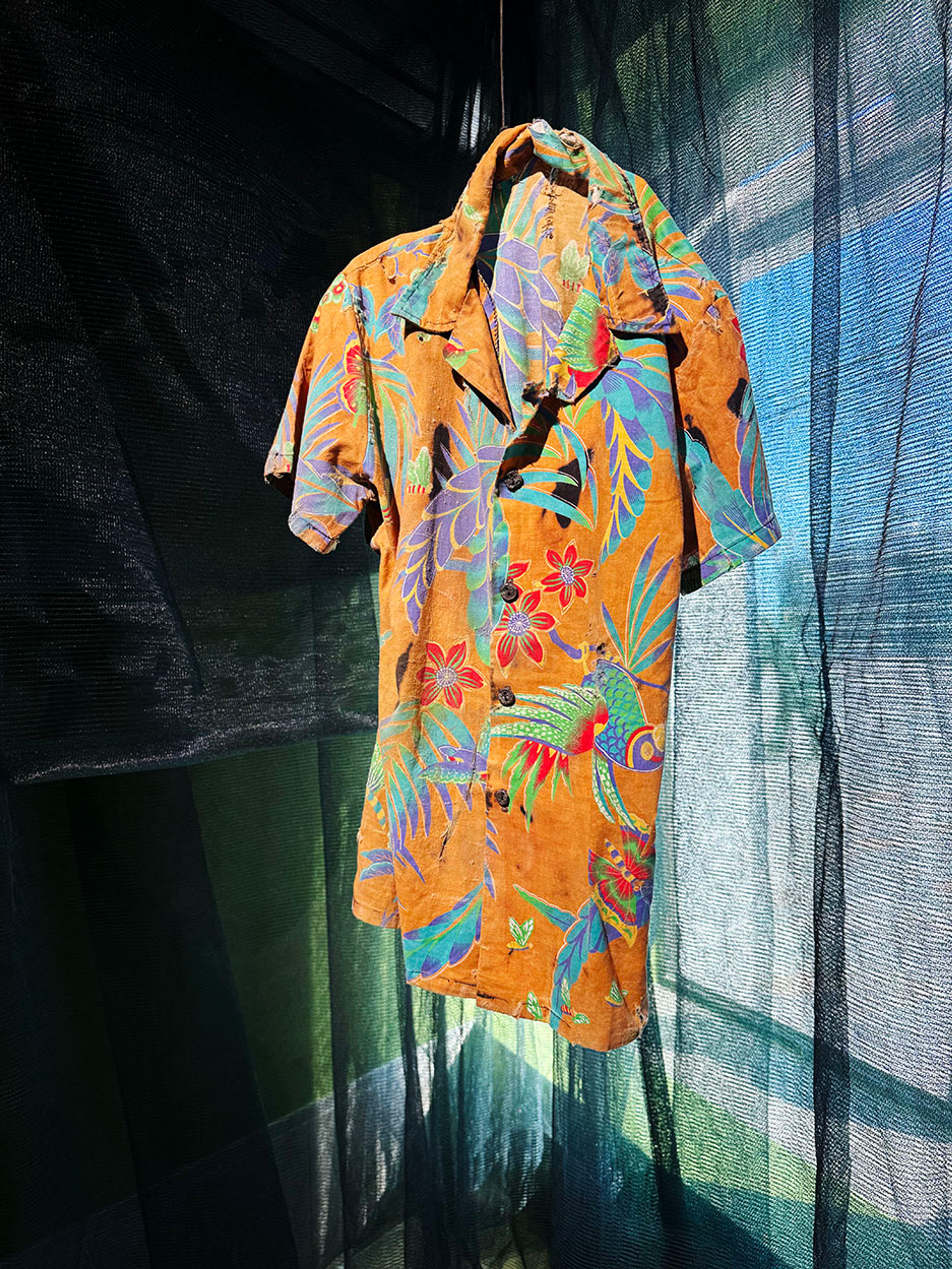 An orange Hawaiian shirt with vibrant tropical patterns hangs against a dark, sheer net.