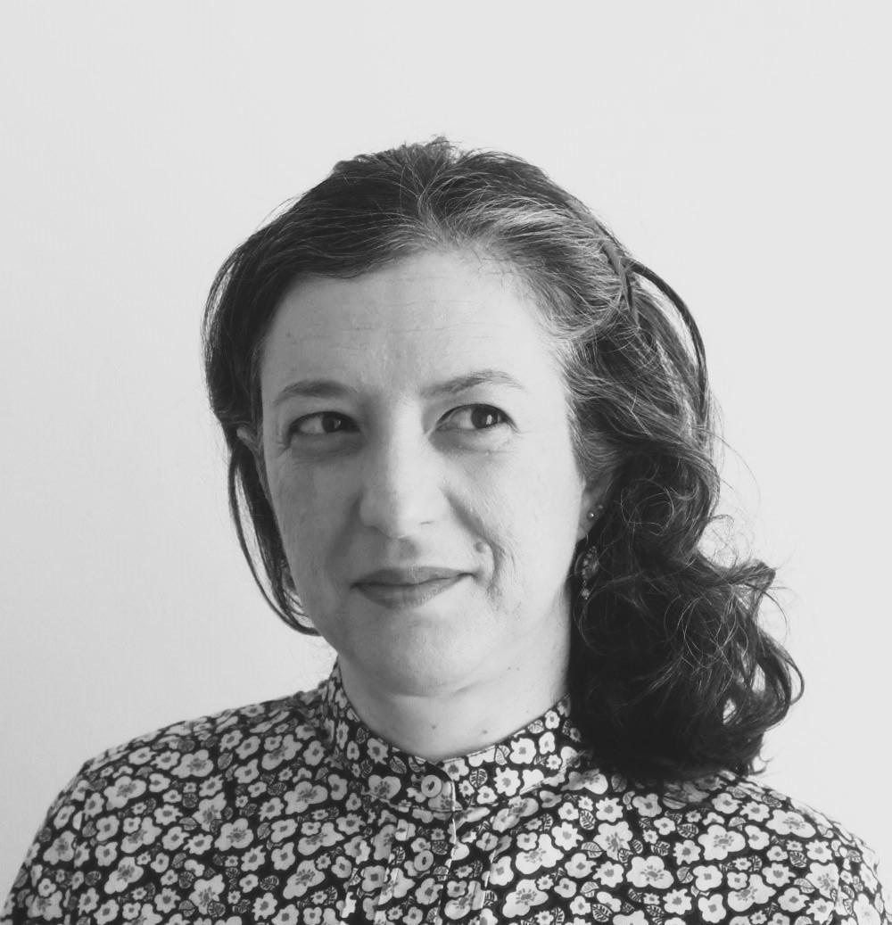 Black and white portrait of a woman with dark, wavy hair, looking to the left with a slight smile, wearing a patterned shirt.