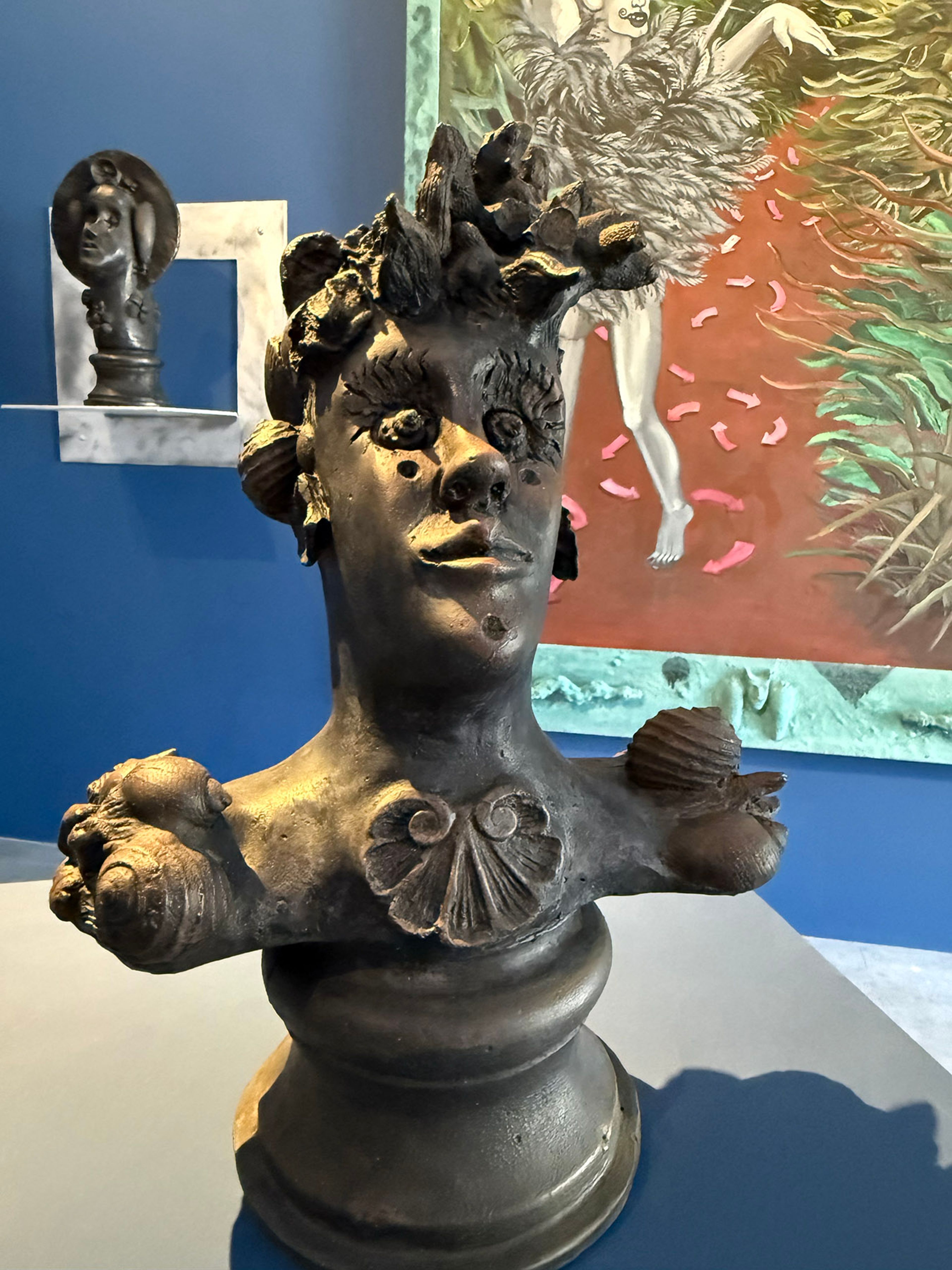 Dark, textured bust of a figure with organic and shell-like details on its head and shoulders, in an art gallery setting.
