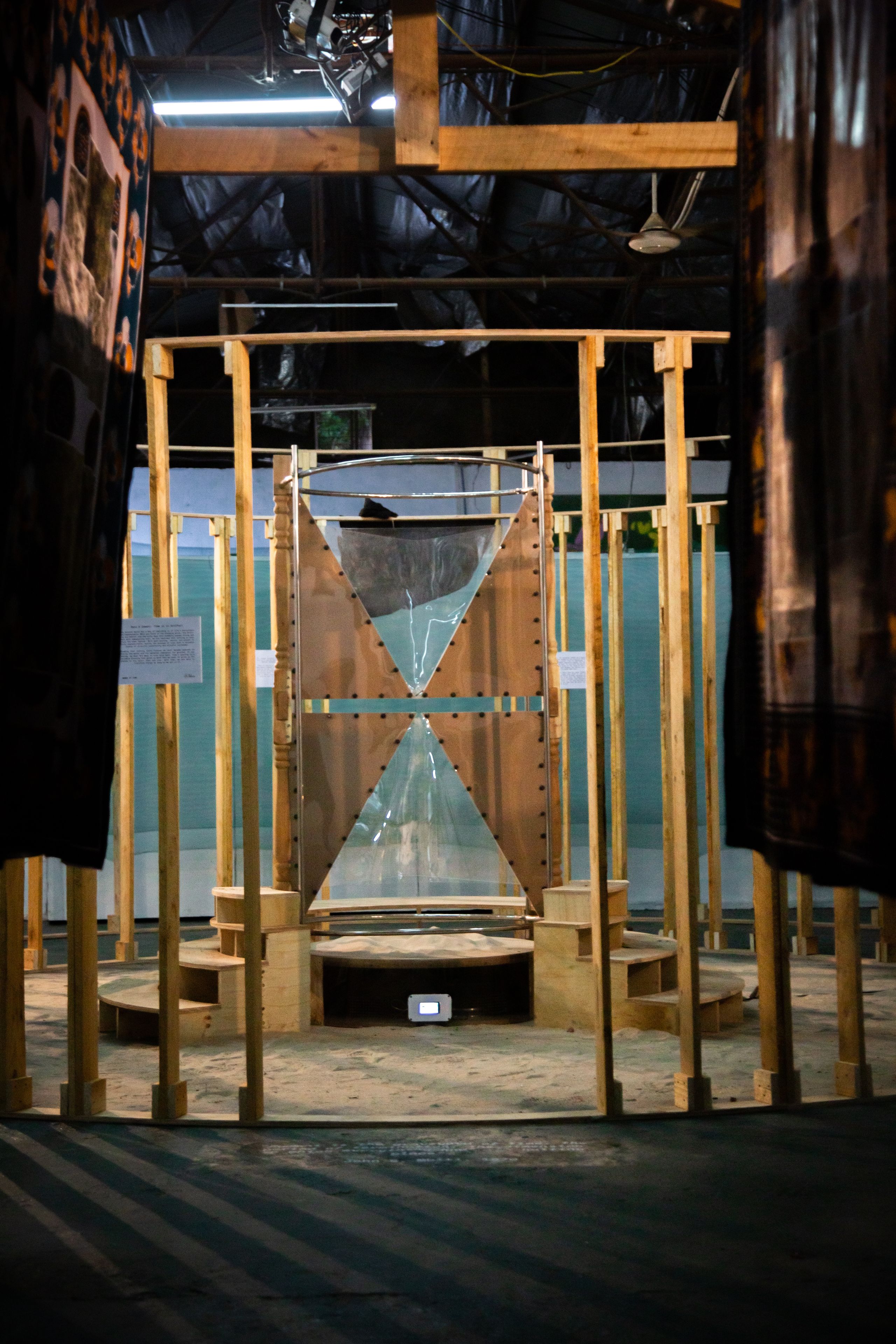 A large hourglass sculpture of wood, metal, and clear panels with sand at its base, within a light wooden framework in a dim industrial space, framed by dark fabric.