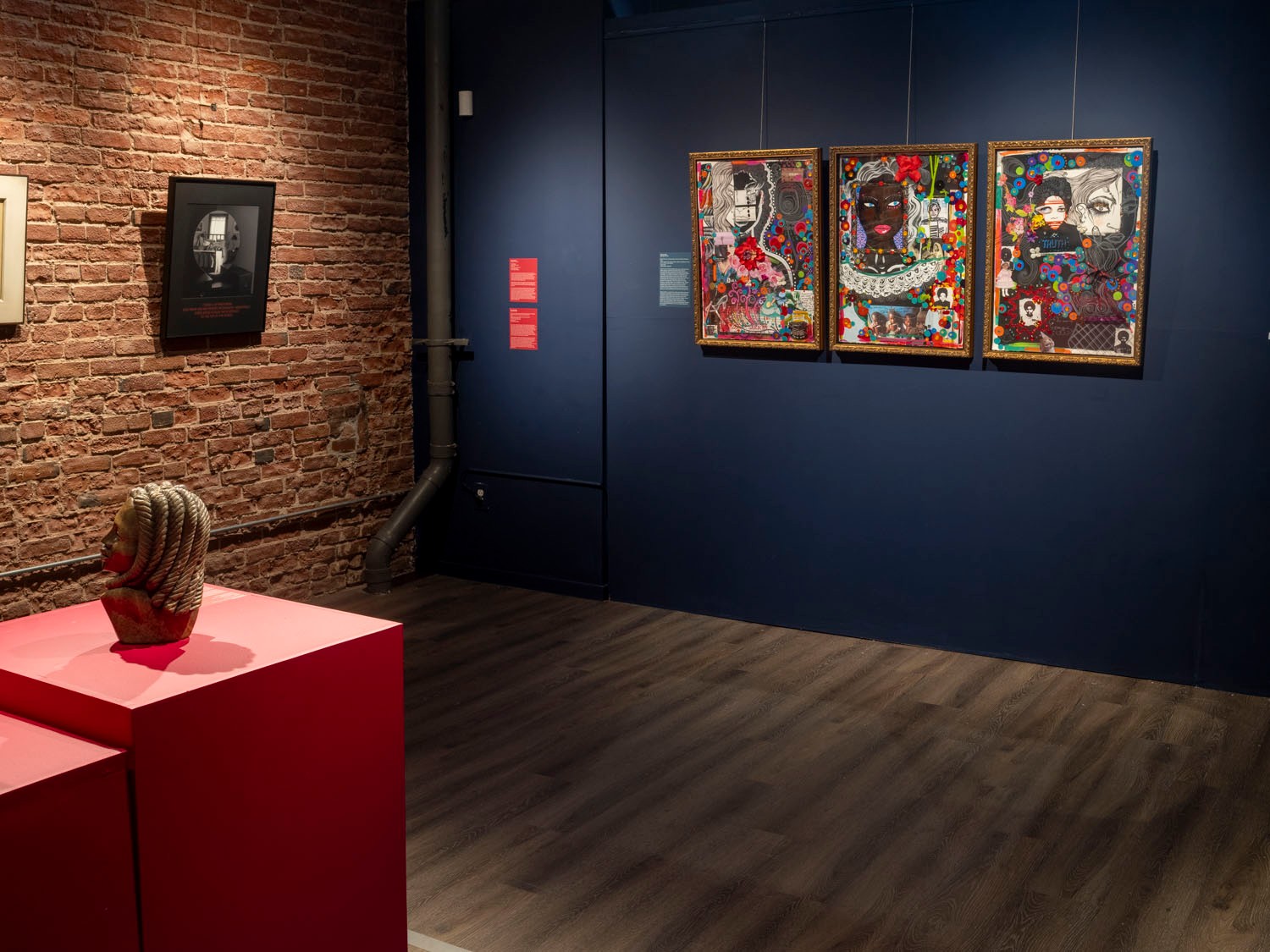 An art gallery featuring a sculpture on red pedestals, a framed photo on a brick wall, and three colorful framed portraits on a dark blue wall.