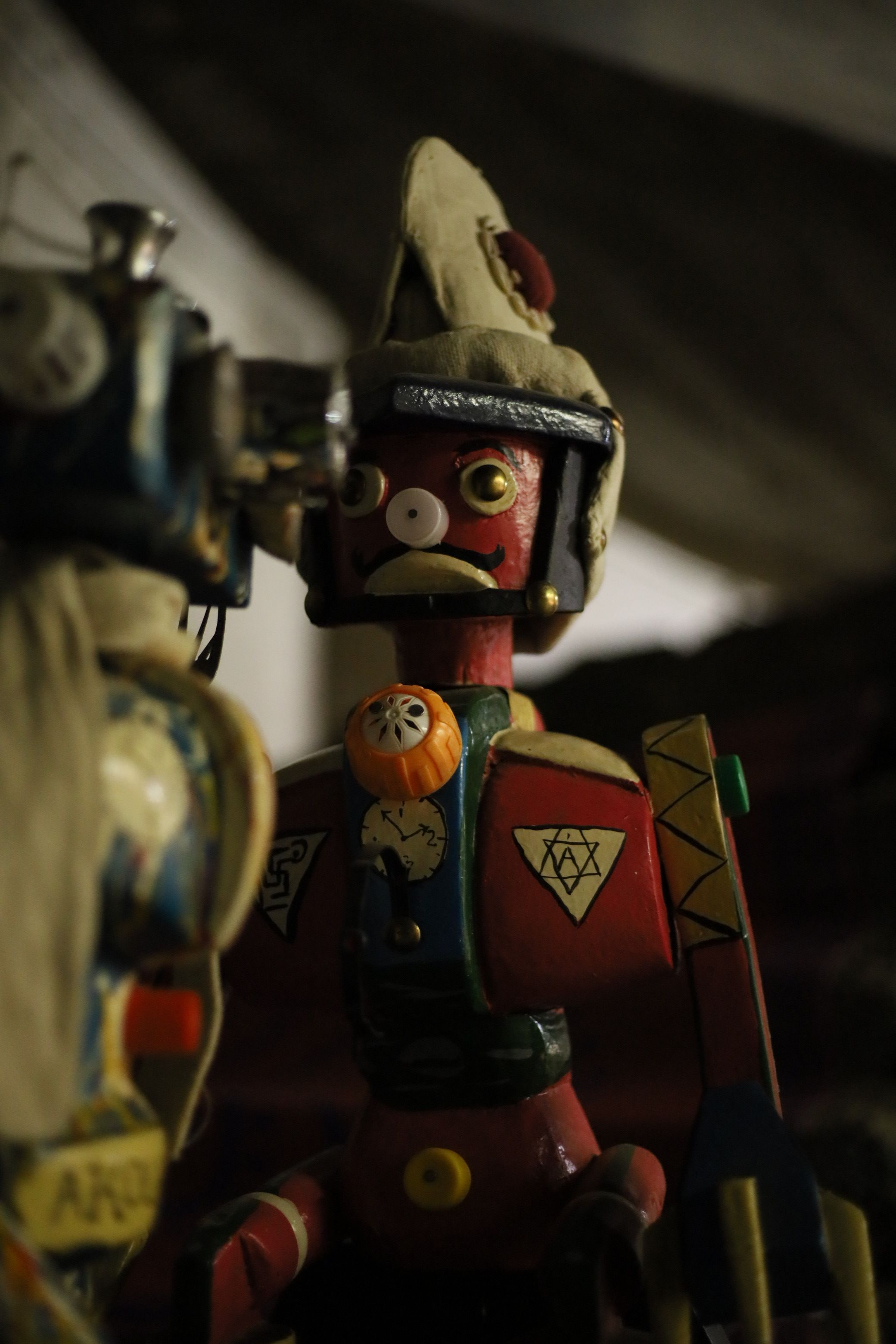 A whimsical, red-faced puppet with a mustache, mismatched button eyes, a beige hat, and a colorful body adorned with a pumpkin charm, clock faces, and symbols, stands in shadow with another puppet blurred on the left.