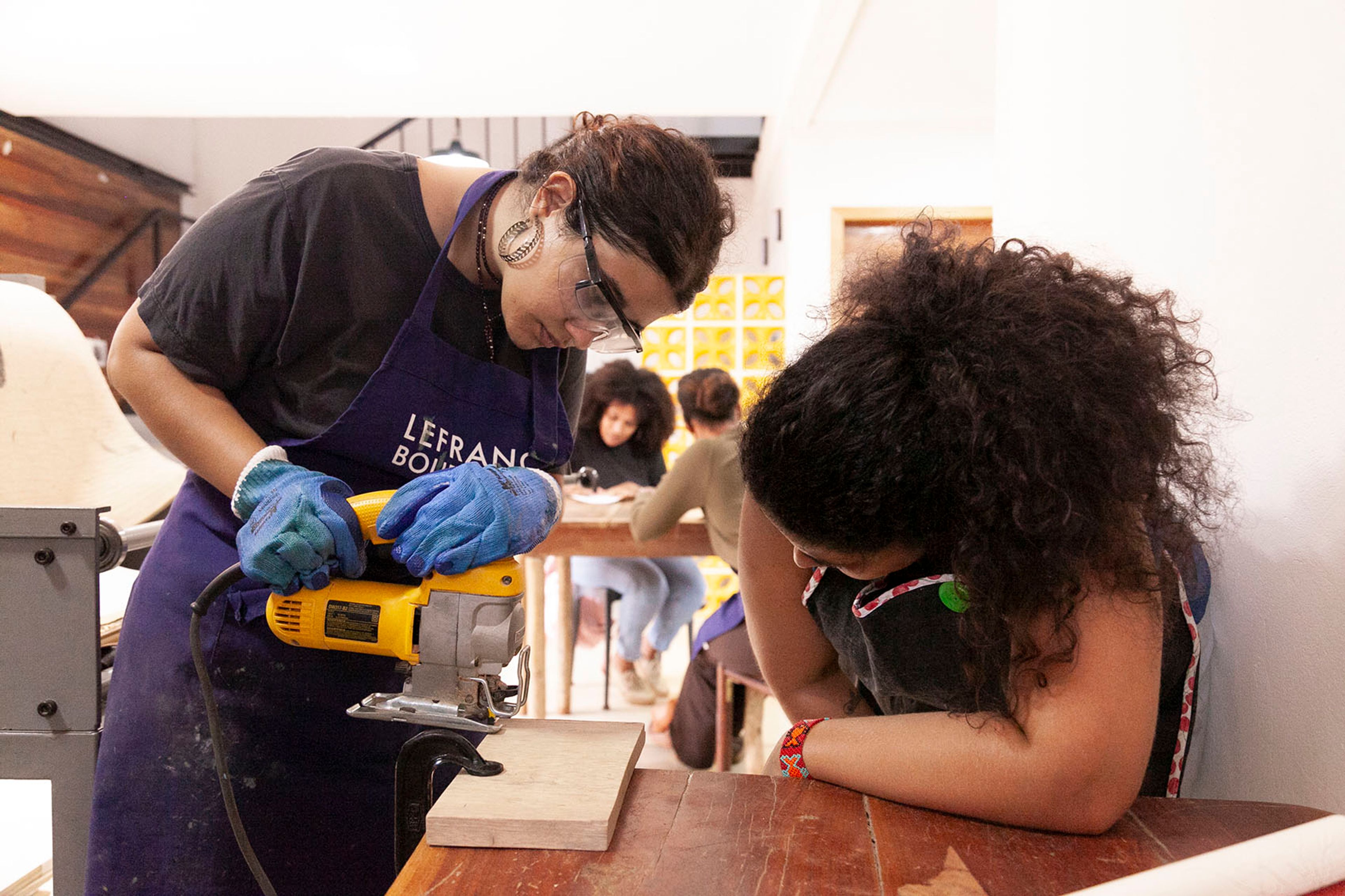 A woman in safety glasses and gloves uses a jigsaw to cut wood, while another woman with curly hair watches in a workshop.