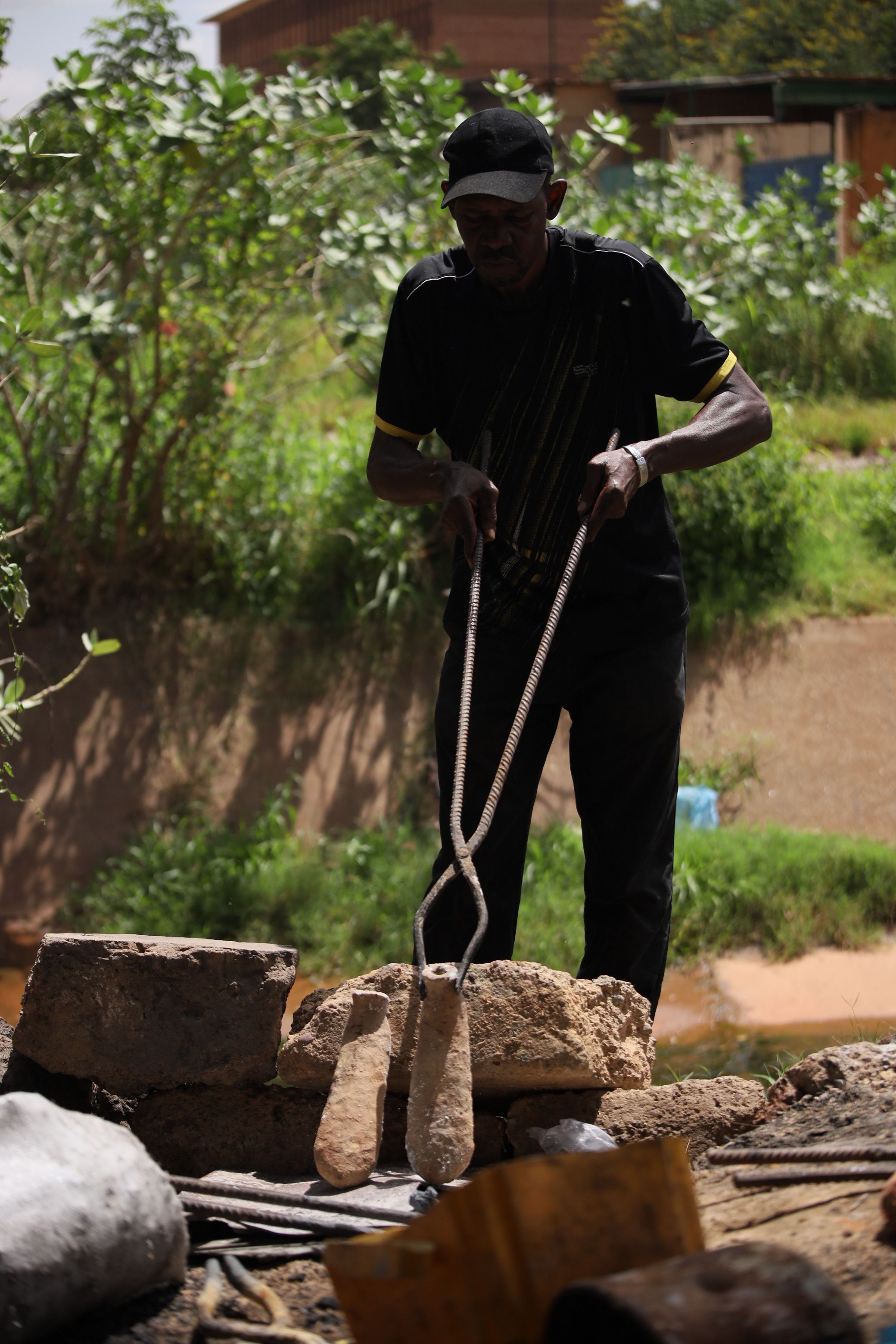 A man in a black cap holds two tapered weights with metal tongs over a stone surface outdoors.