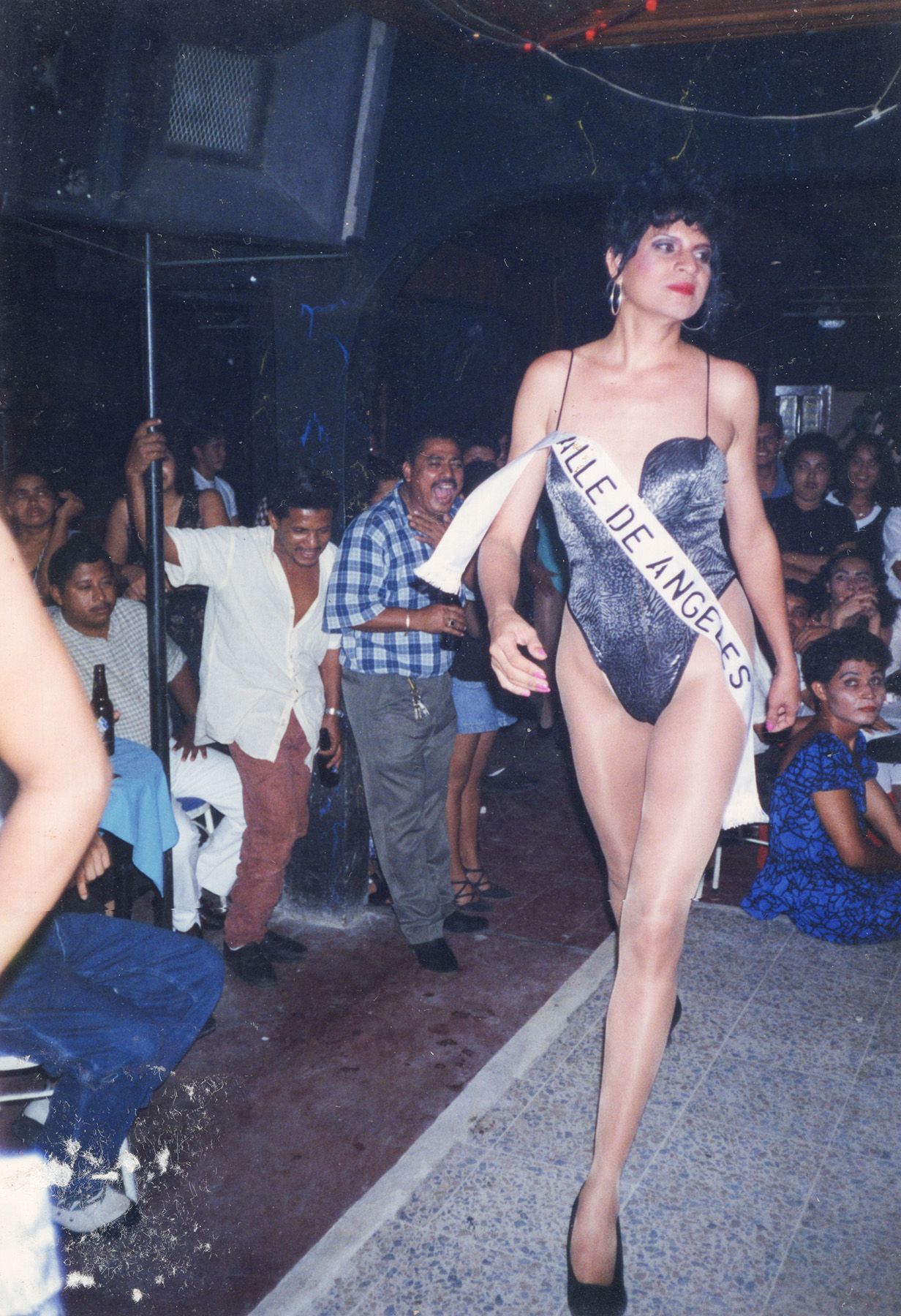 A pageant contestant in a sparkly gray swimsuit and "Valle de Angeles" sash walks a dimly lit runway past an excited audience.