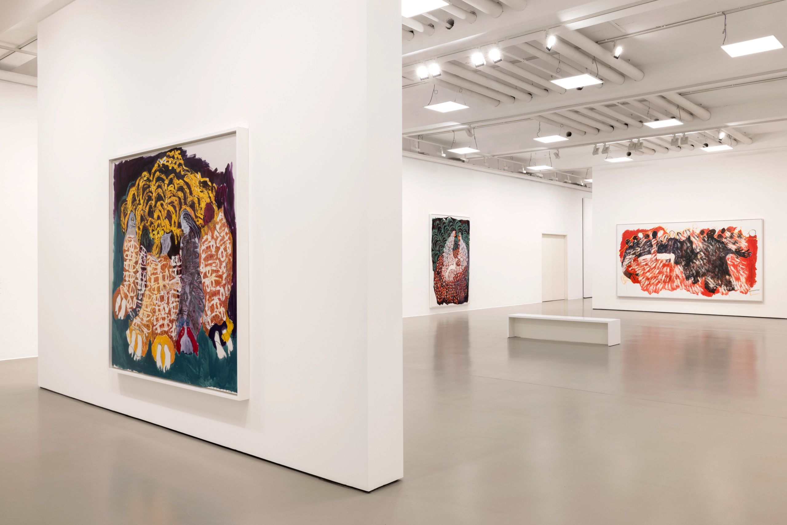 A modern art gallery with white walls and a grey floor, featuring several large, abstract paintings. A vibrant, colorful piece is prominent on a foreground wall, while a large red and black abstract painting hangs in the background, near a white bench.