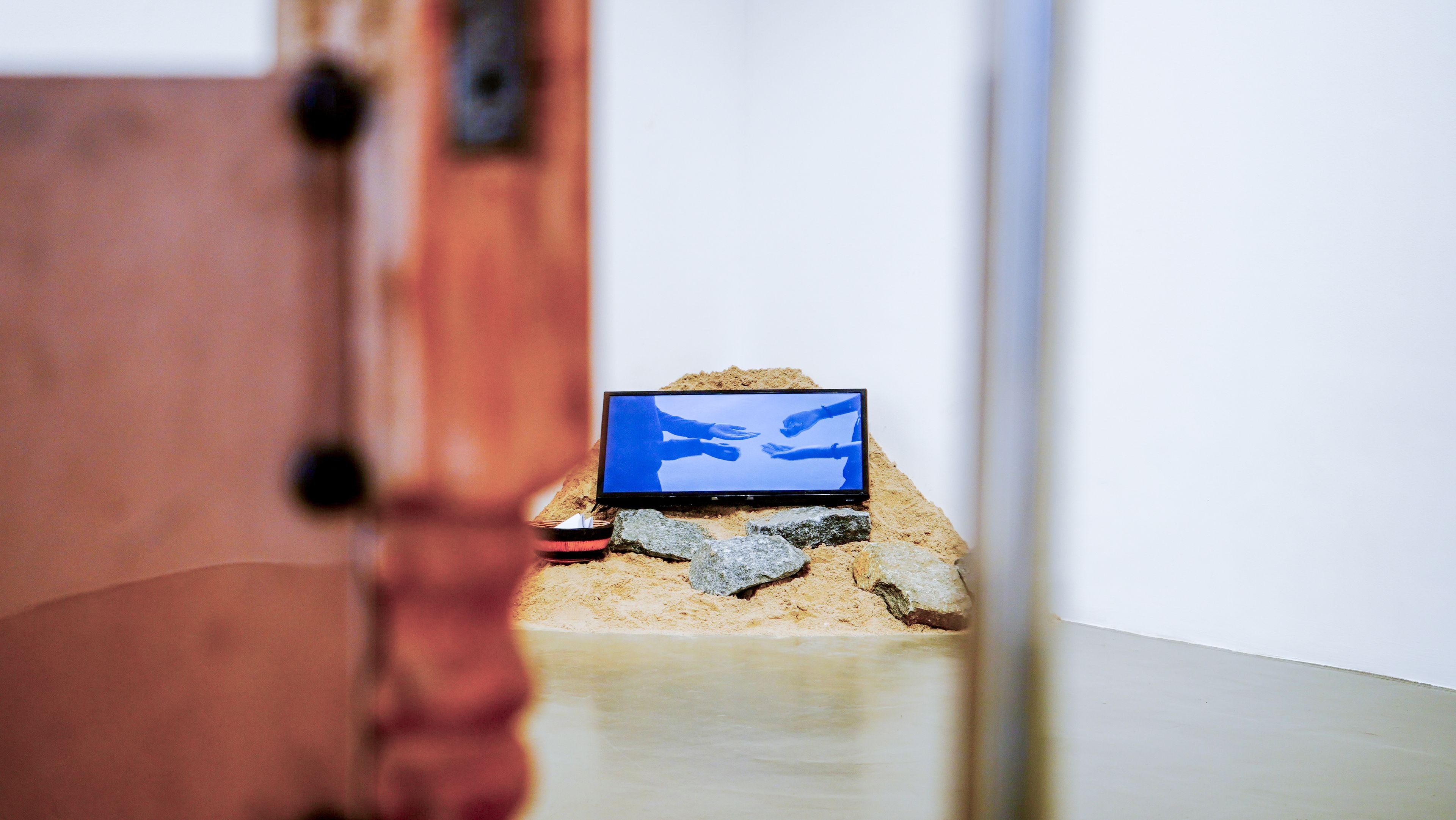A monitor displaying blue hands is set on a pile of sand and rocks in a gallery.