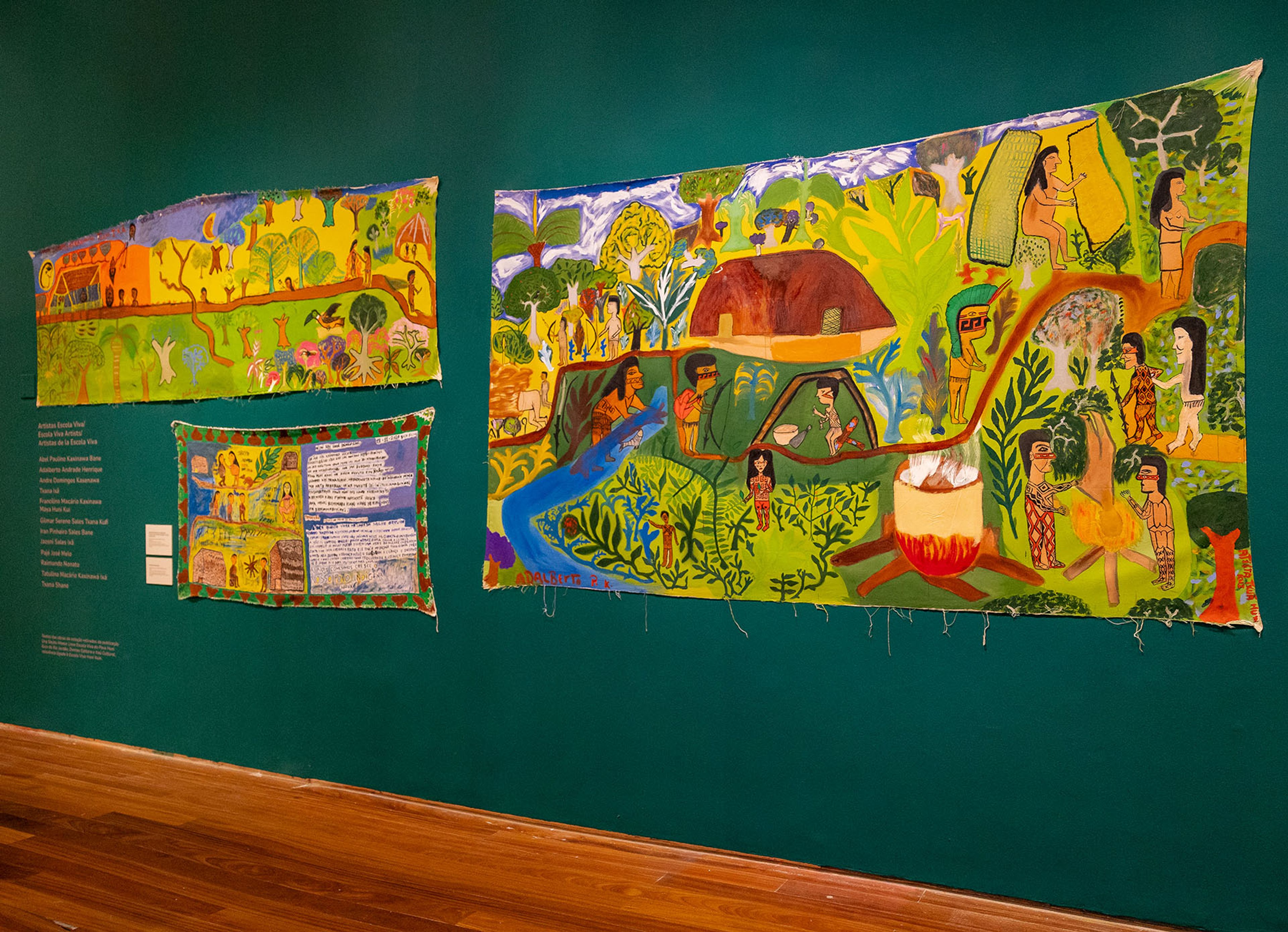 Three colorful folk art textile artworks depicting indigenous people and jungle scenes hang on a green museum wall above a wooden floor.