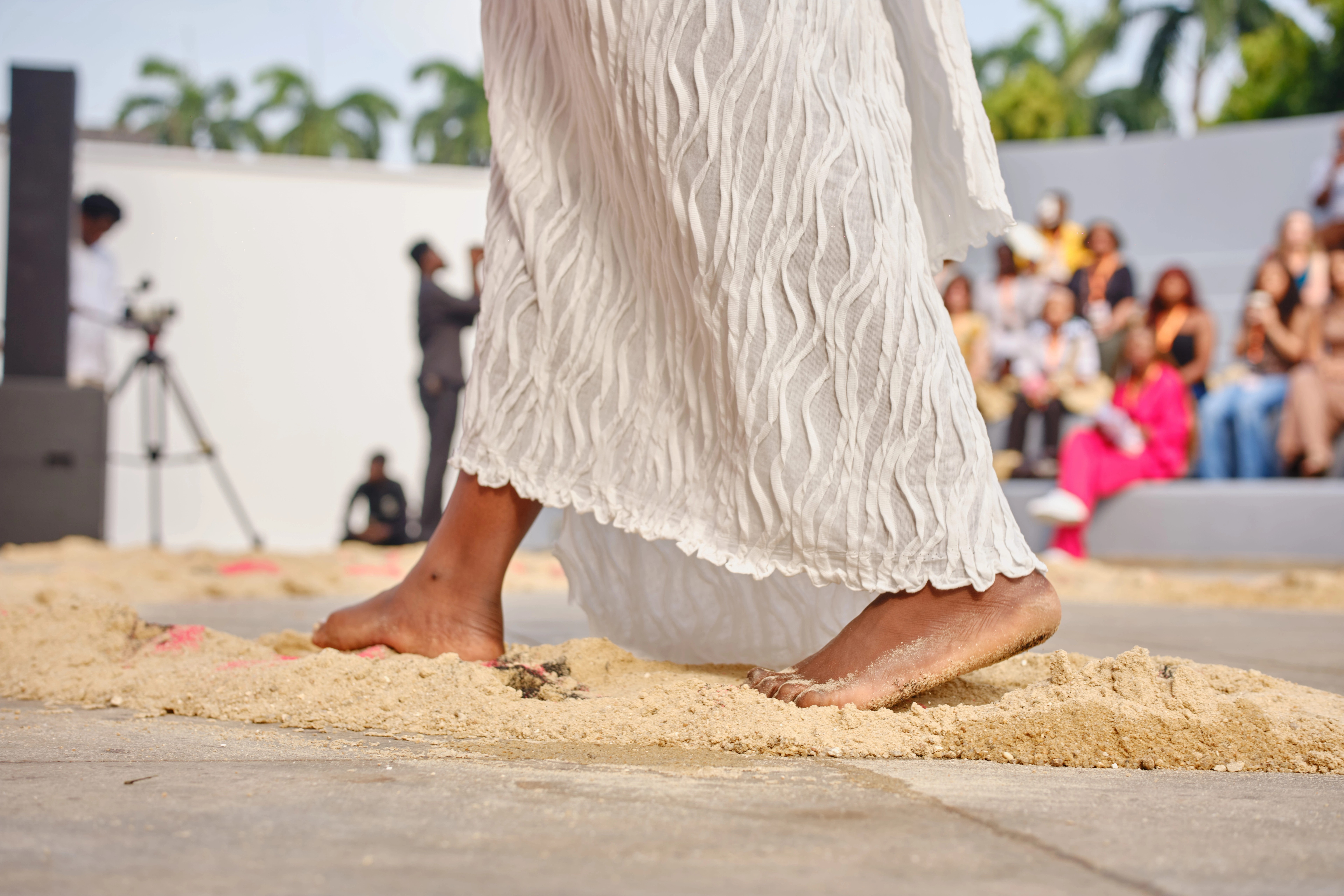 Bare feet in a white dress stepping on a sandy path, with a blurred audience in the background.