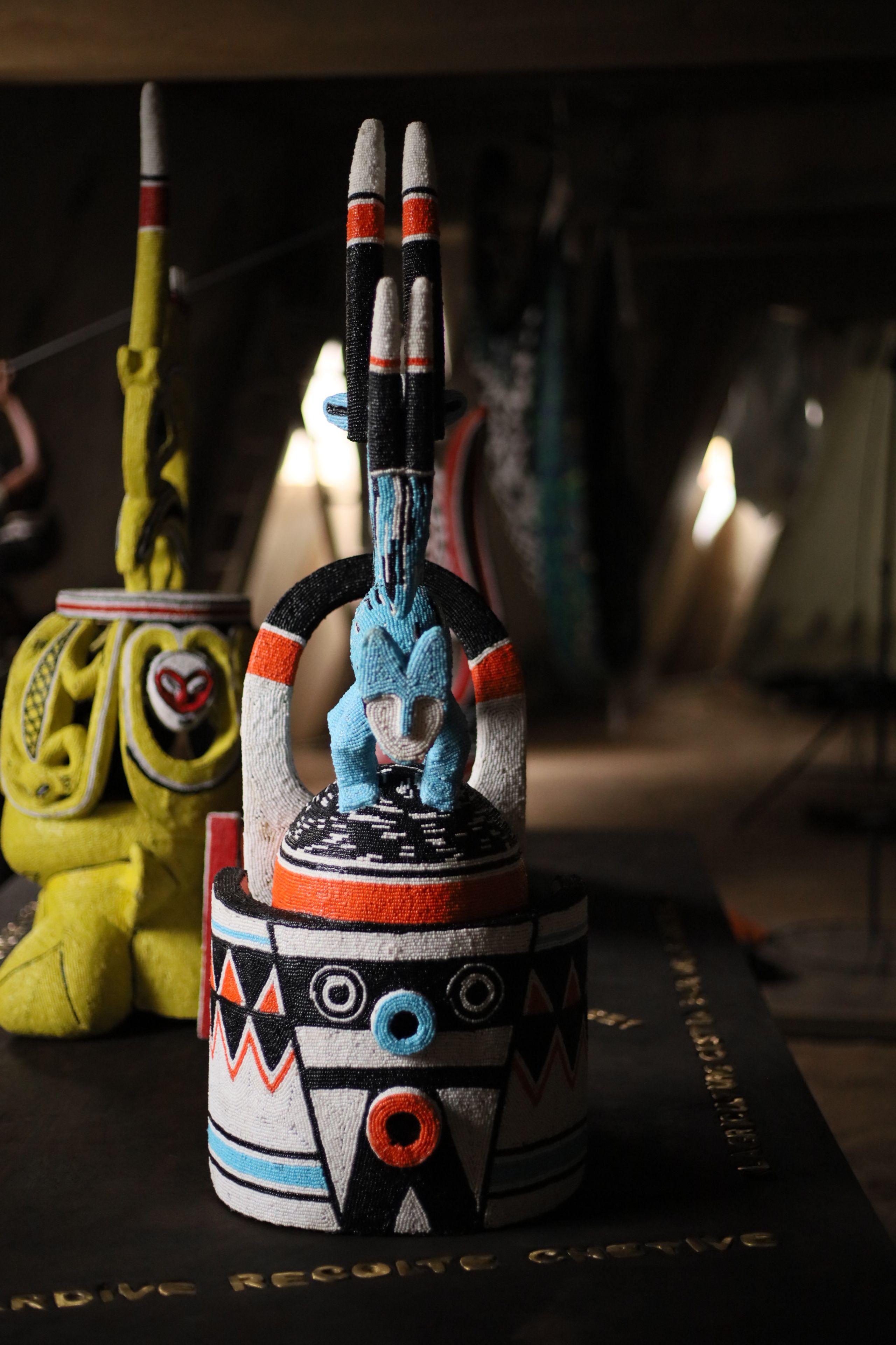 Beaded sculpture with a blue fox-like figure, geometric patterns in black, white, orange, and blue, and four pointed spires. Another yellow sculpture is blurred behind it.