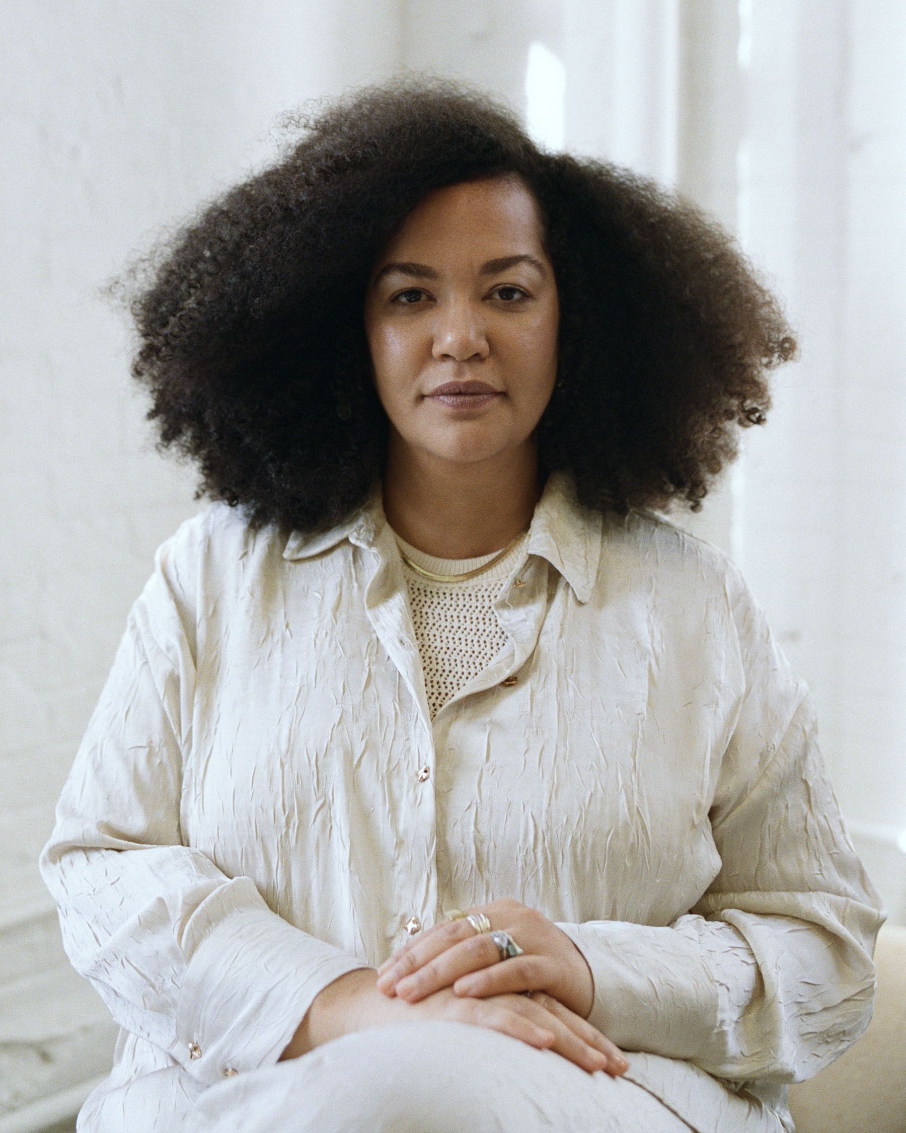 A woman with voluminous dark curly hair, wearing a cream textured shirt, clasps her hands and looks straight ahead.