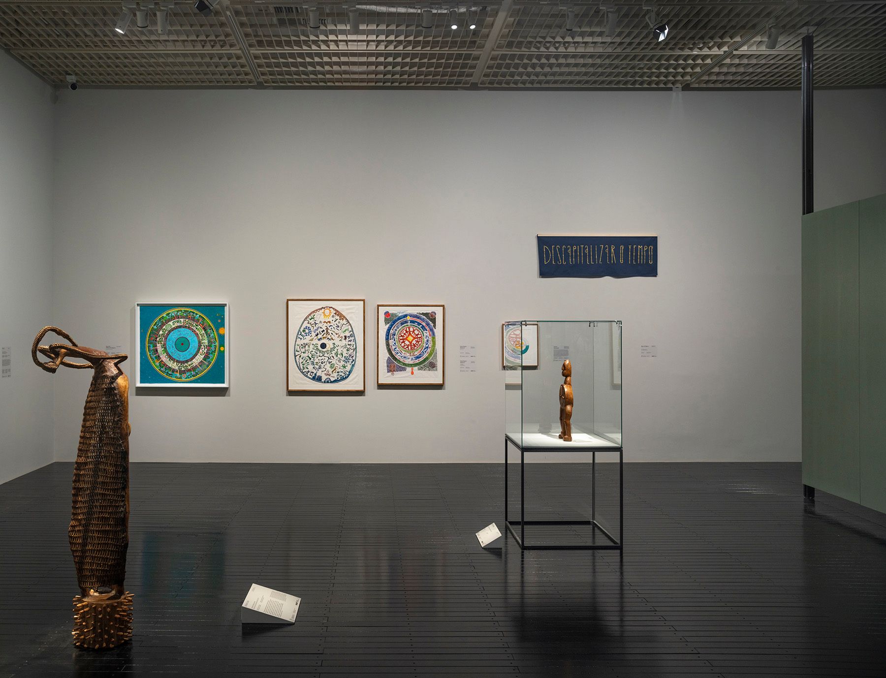 An art gallery displays a tall wooden sculpture, three colorful framed artworks, a small wooden figure in a glass case, and a blue banner with text on the wall.