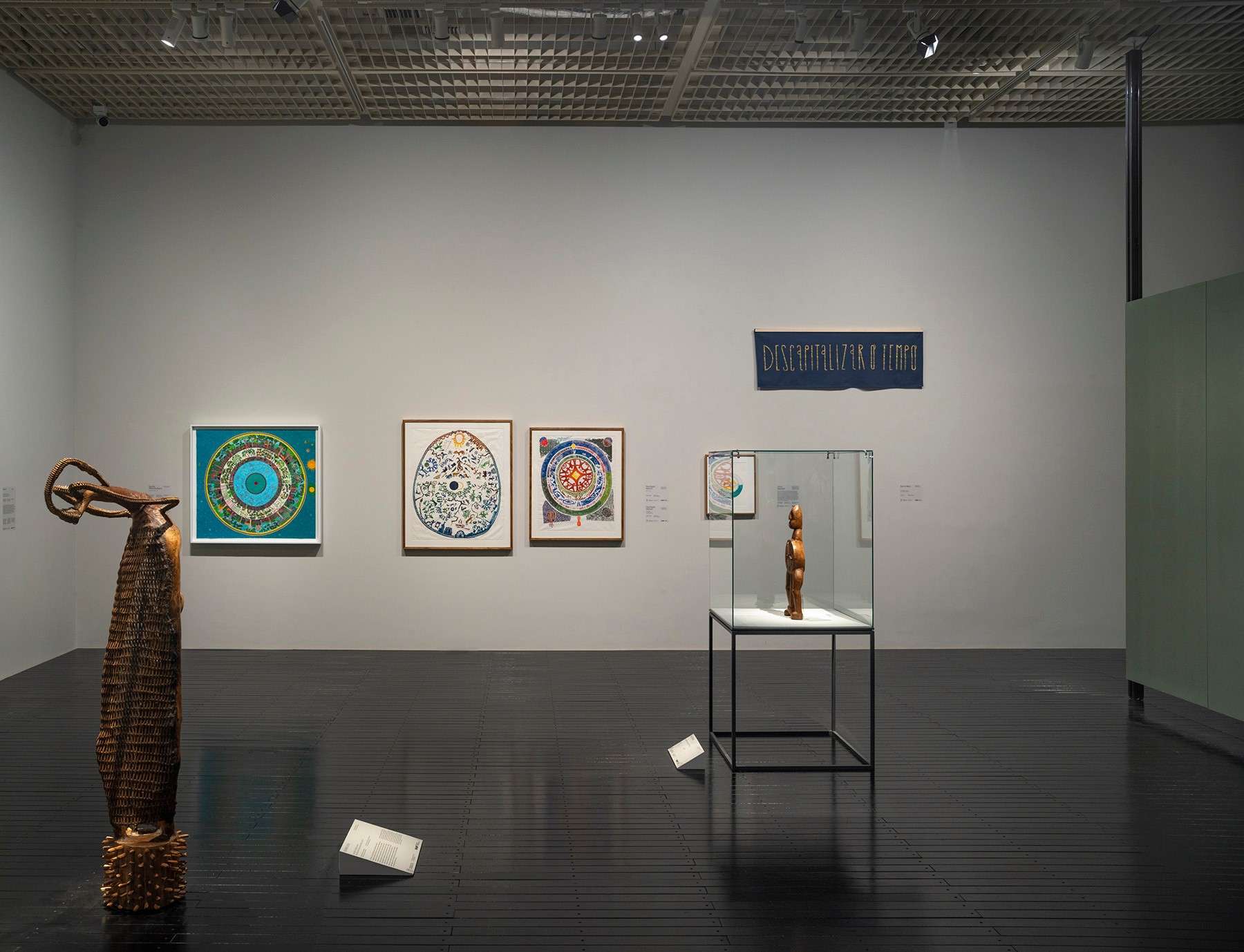 An art gallery displays a tall wooden sculpture, three colorful framed artworks, a small wooden figure in a glass case, and a blue banner with text on the wall.