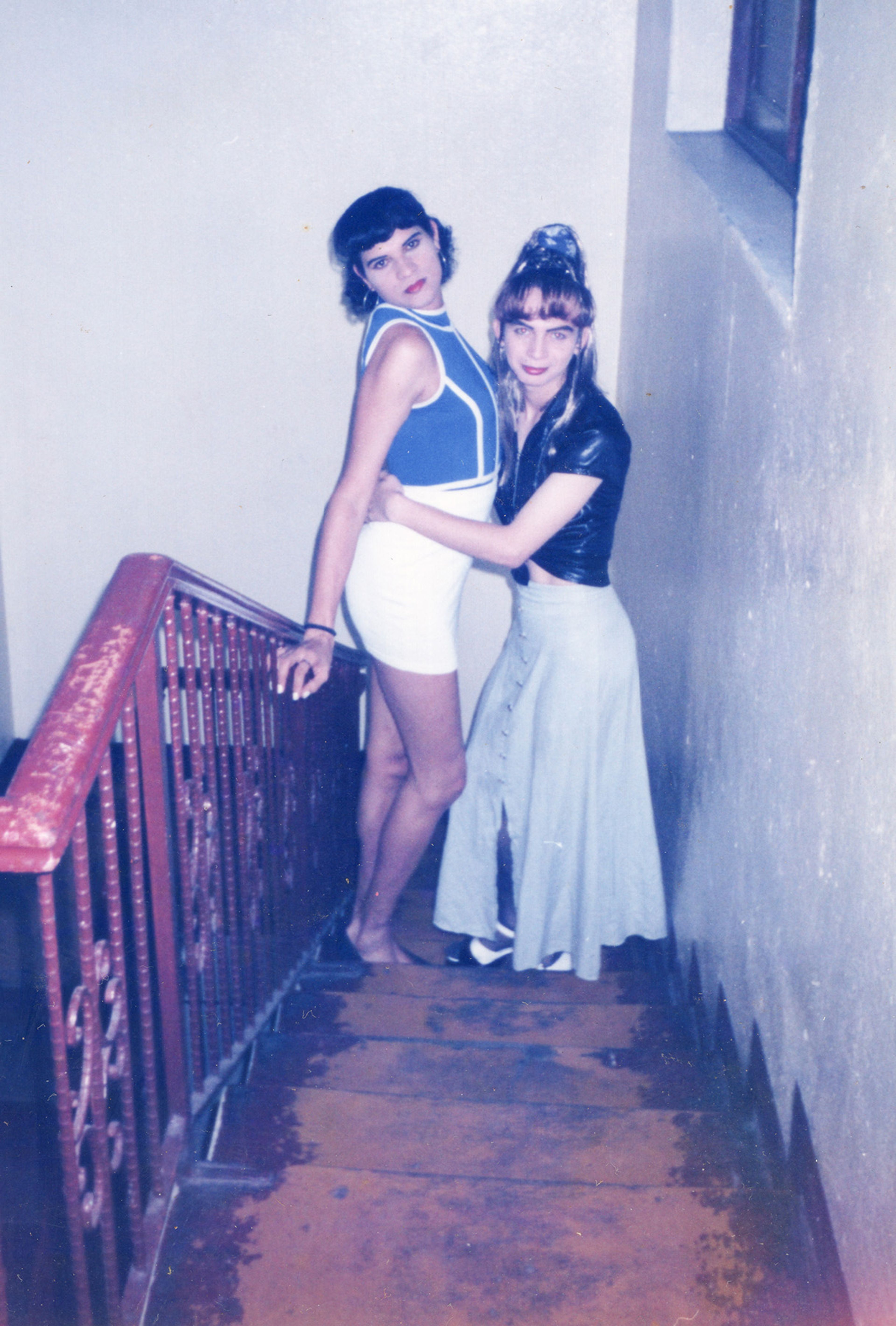 Two individuals in vintage clothing pose together on a wooden staircase, one embracing the other.