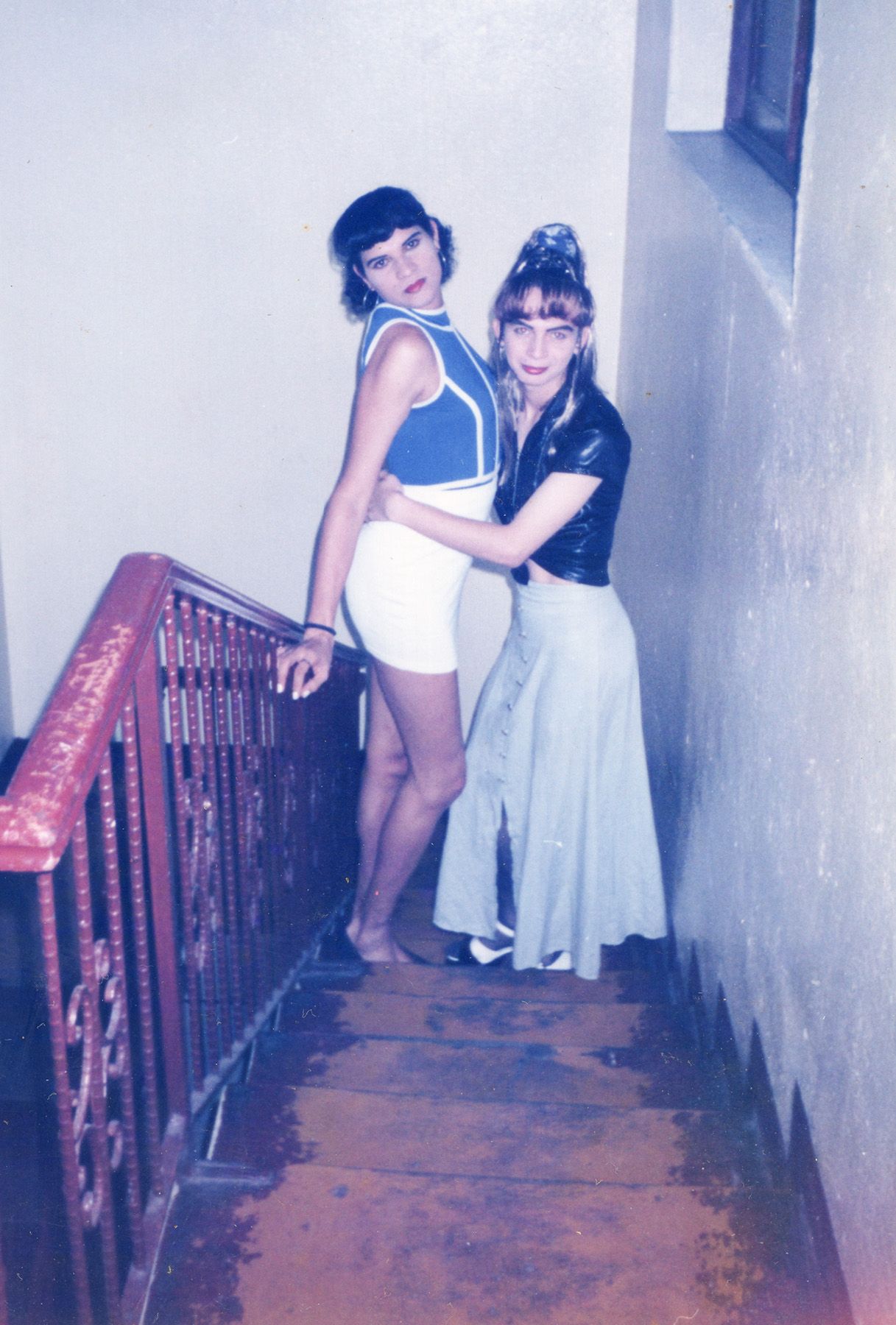 Two individuals in vintage clothing pose together on a wooden staircase, one embracing the other.