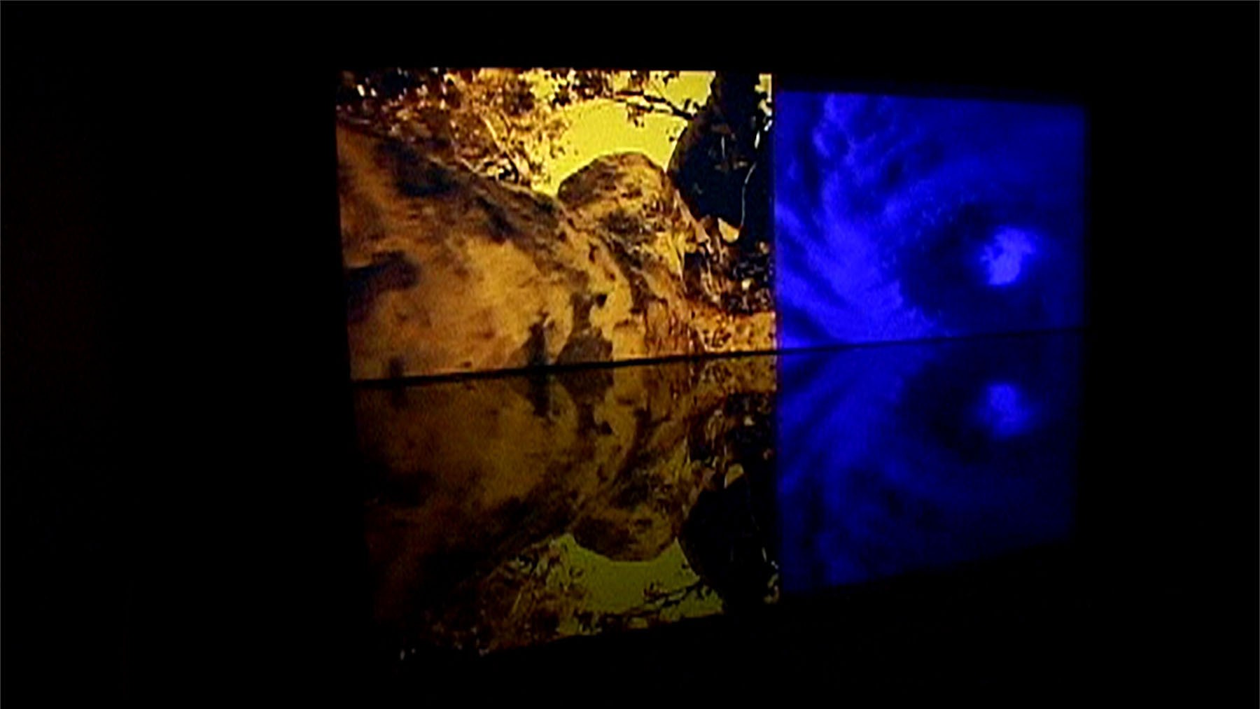 Two projected panels: a yellow-brown abstract form and a blue swirling hurricane, both reflected on a dark surface.