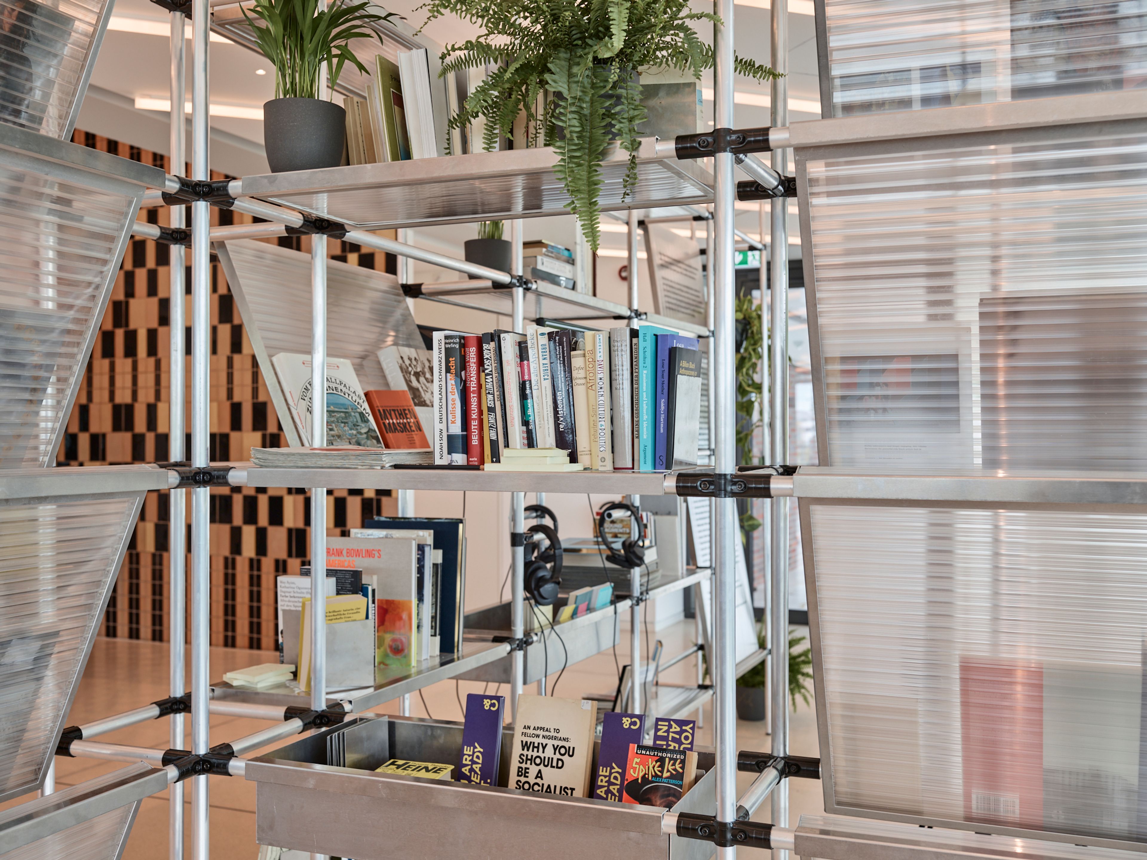 A modern modular shelving unit with metal poles, translucent panels, books, and potted plants.