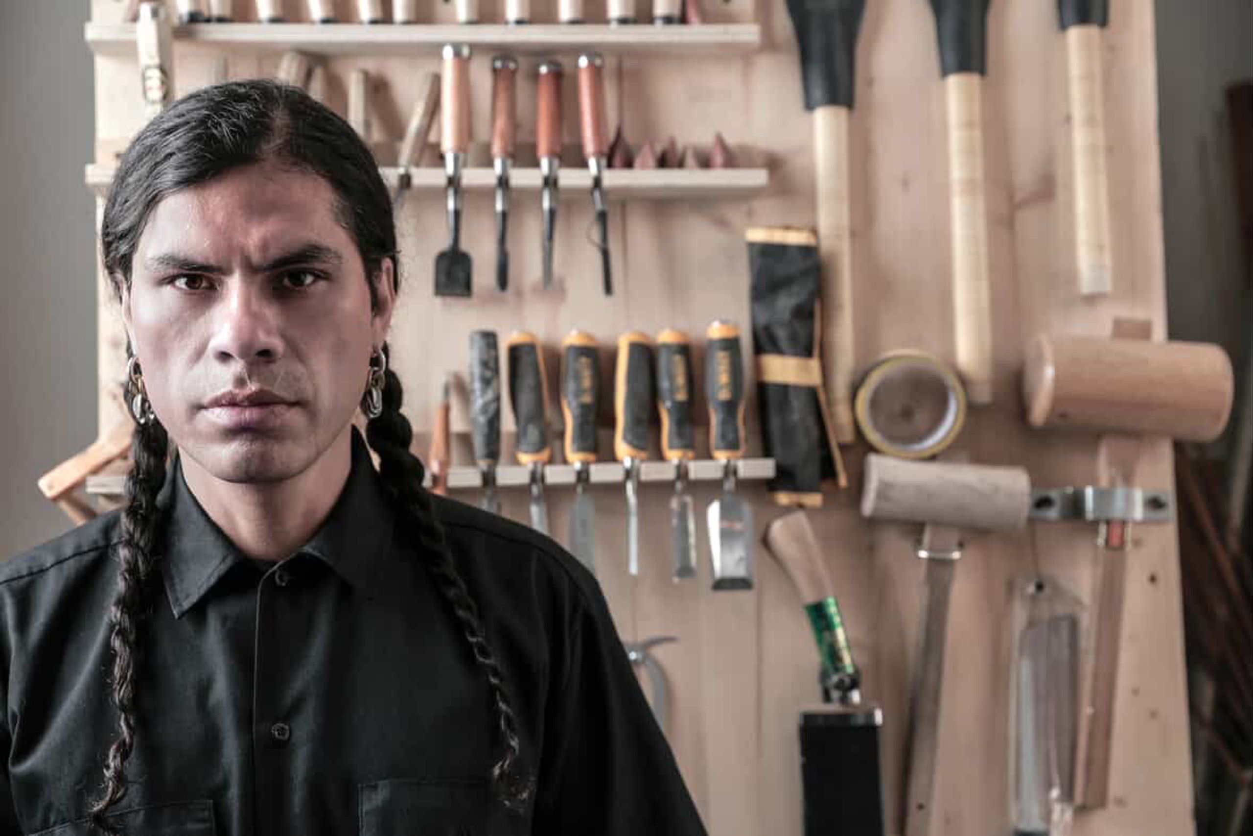 A man with long braided hair and earrings looks seriously at the camera in front of a wall of woodworking tools.