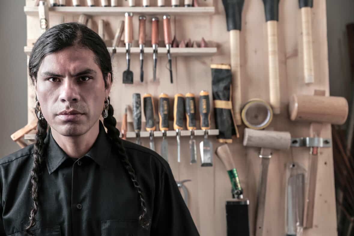 A man with long braided hair and earrings looks seriously at the camera in front of a wall of woodworking tools.