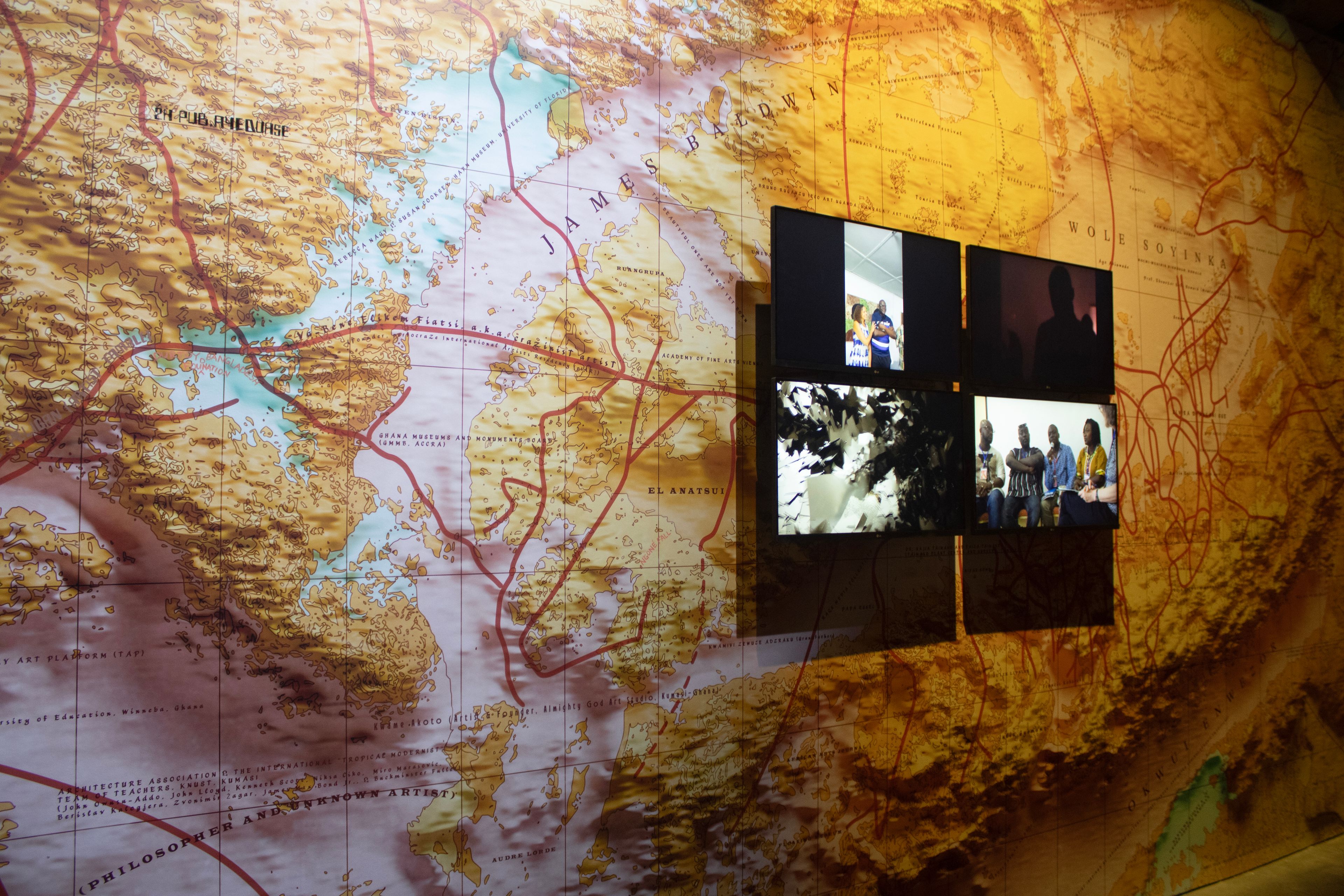 Four screens displaying various videos are mounted on a large, textured wall map.