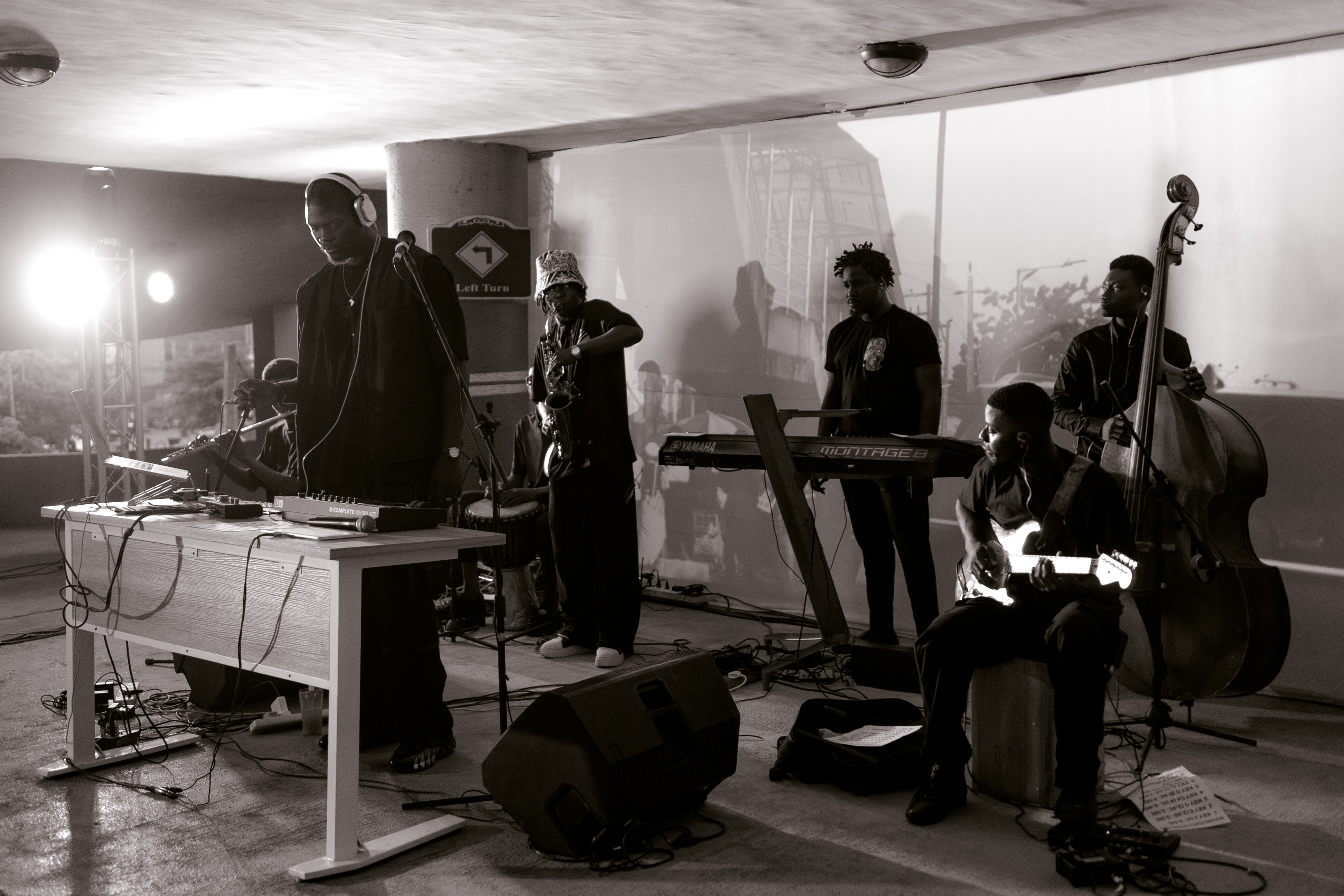 Black and white photo of a band performing, with a DJ, upright bassist, keyboardist, and seated guitarist visible.