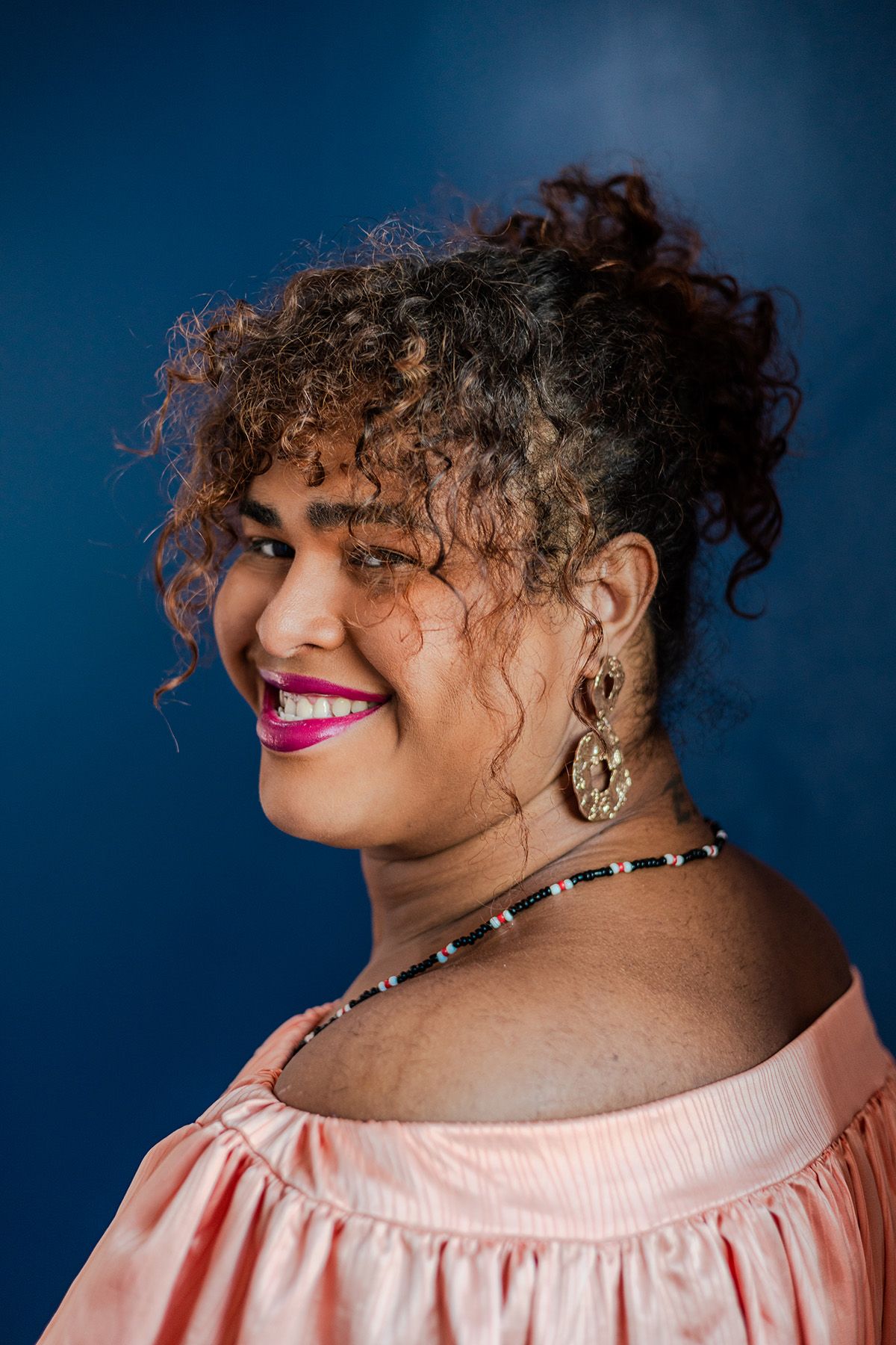 Smiling woman with curly hair and pink lipstick, wearing a peach off-shoulder top, gold earrings, and a beaded necklace, looking over her shoulder against a blue background.