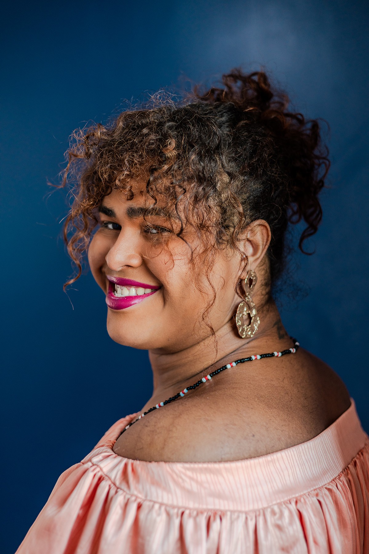 Smiling woman with curly hair and pink lipstick, wearing a peach off-shoulder top, gold earrings, and a beaded necklace, looking over her shoulder against a blue background.