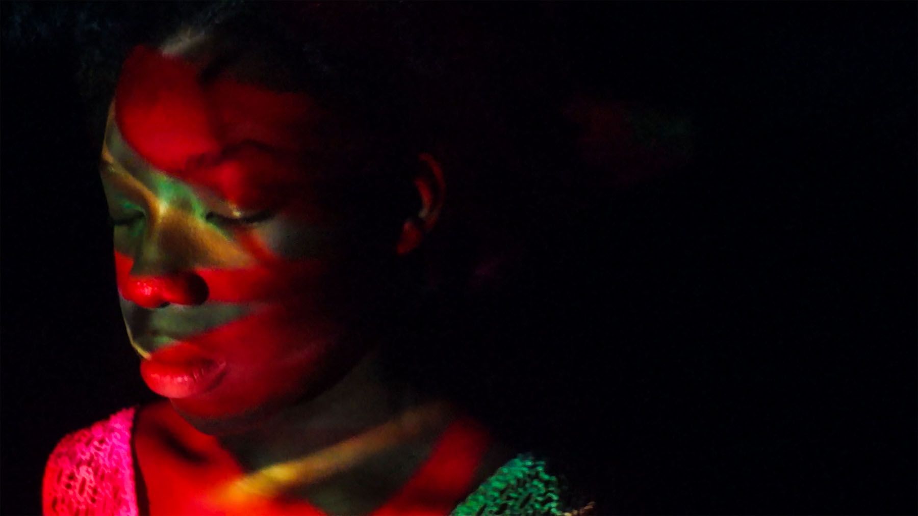 A dark-skinned person with closed eyes, their face and shoulder illuminated by red and green light patterns.