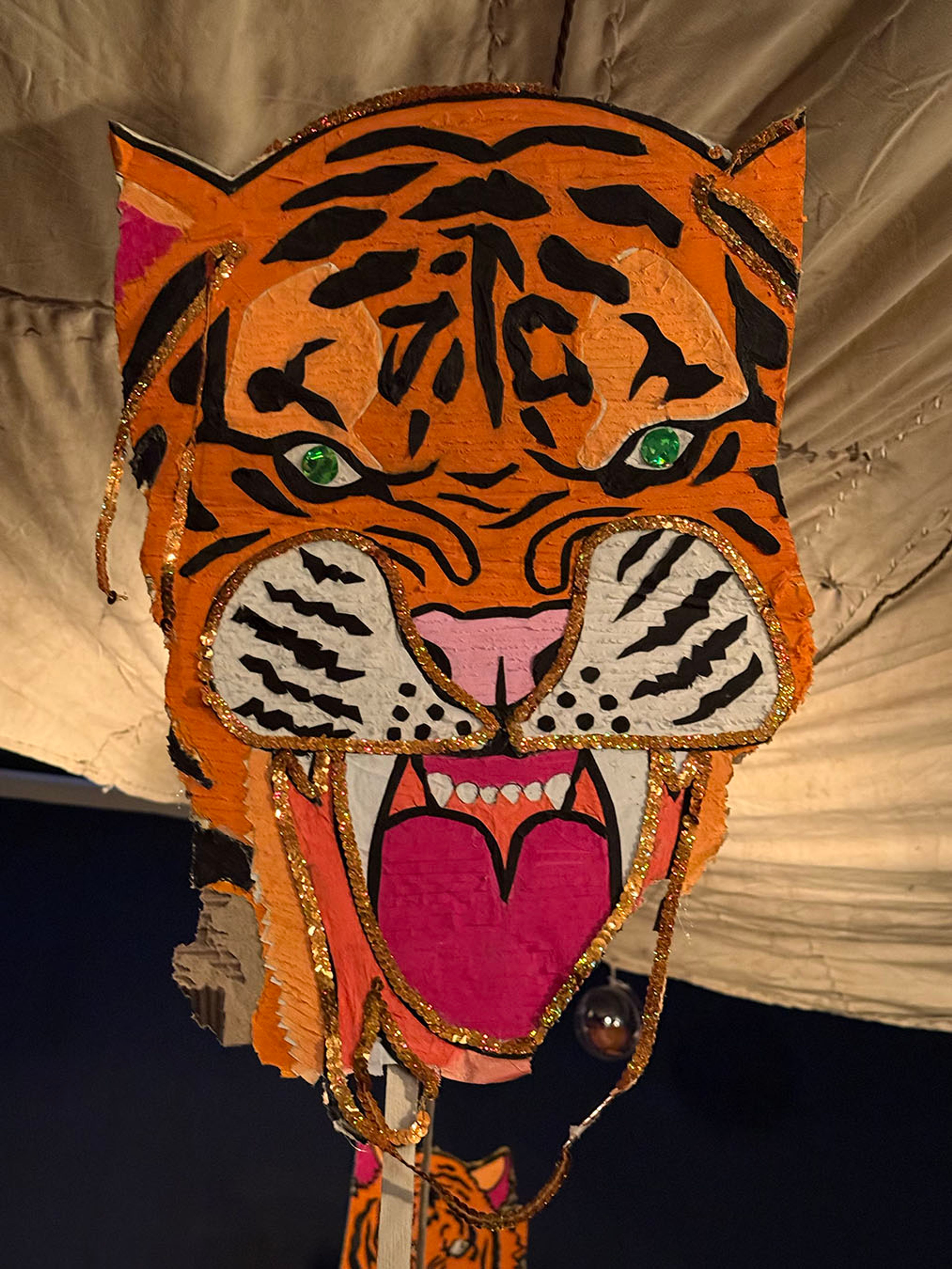 An orange and black tiger head decoration with green eyes, a pink open mouth, and gold glitter accents.