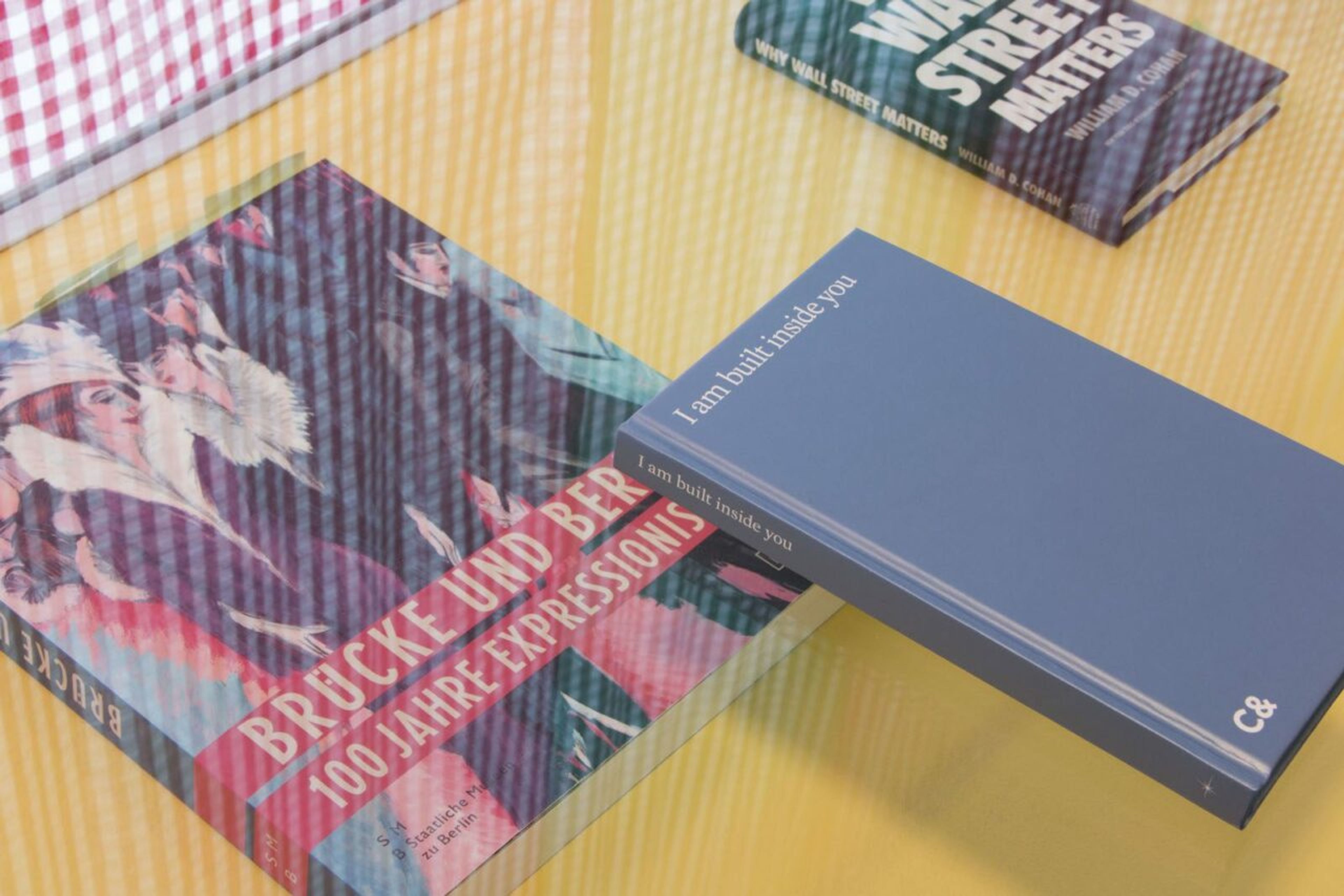 Three books on a yellow surface: an art book with a colorful figurative cover, a blue book titled "I am built inside you", and a book titled "WHY WALL STREET MATTERS".
