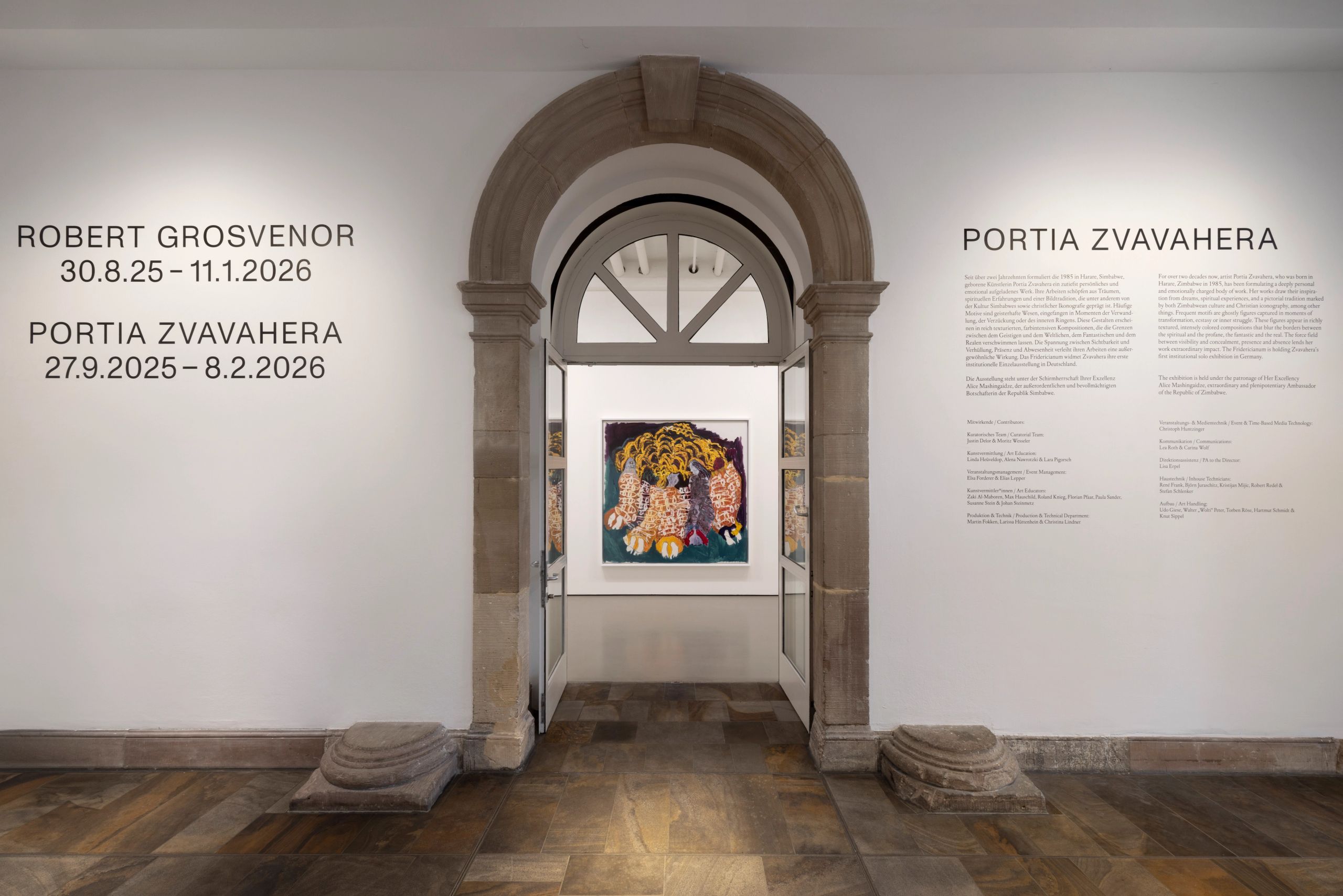 A gallery wall displaying exhibition titles and dates on the left, and a description for "Portia Zvavahera" on the right, framing a stone archway through which a colorful painting is visible in the next room.