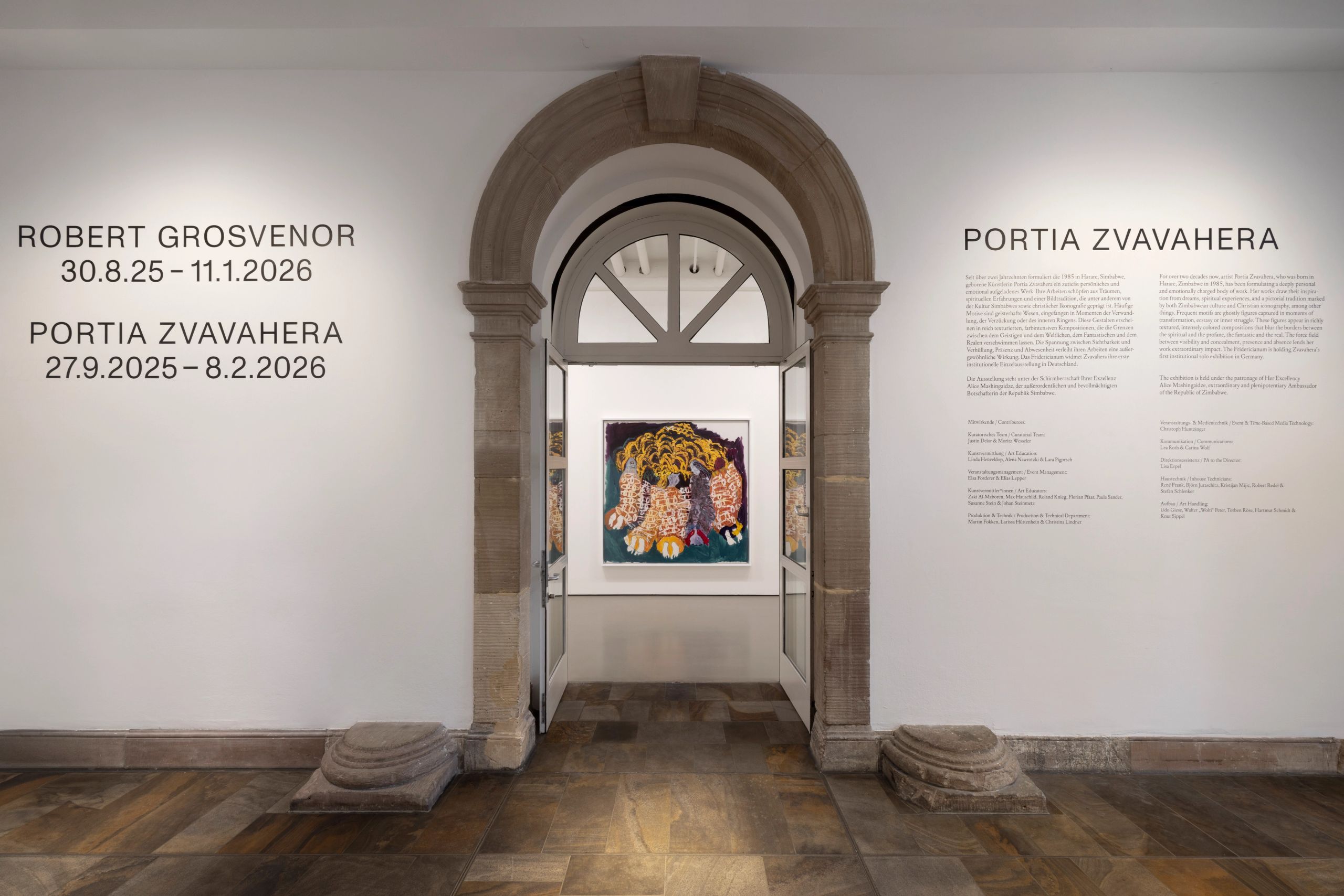 A gallery wall displaying exhibition titles and dates on the left, and a description for "Portia Zvavahera" on the right, framing a stone archway through which a colorful painting is visible in the next room.
