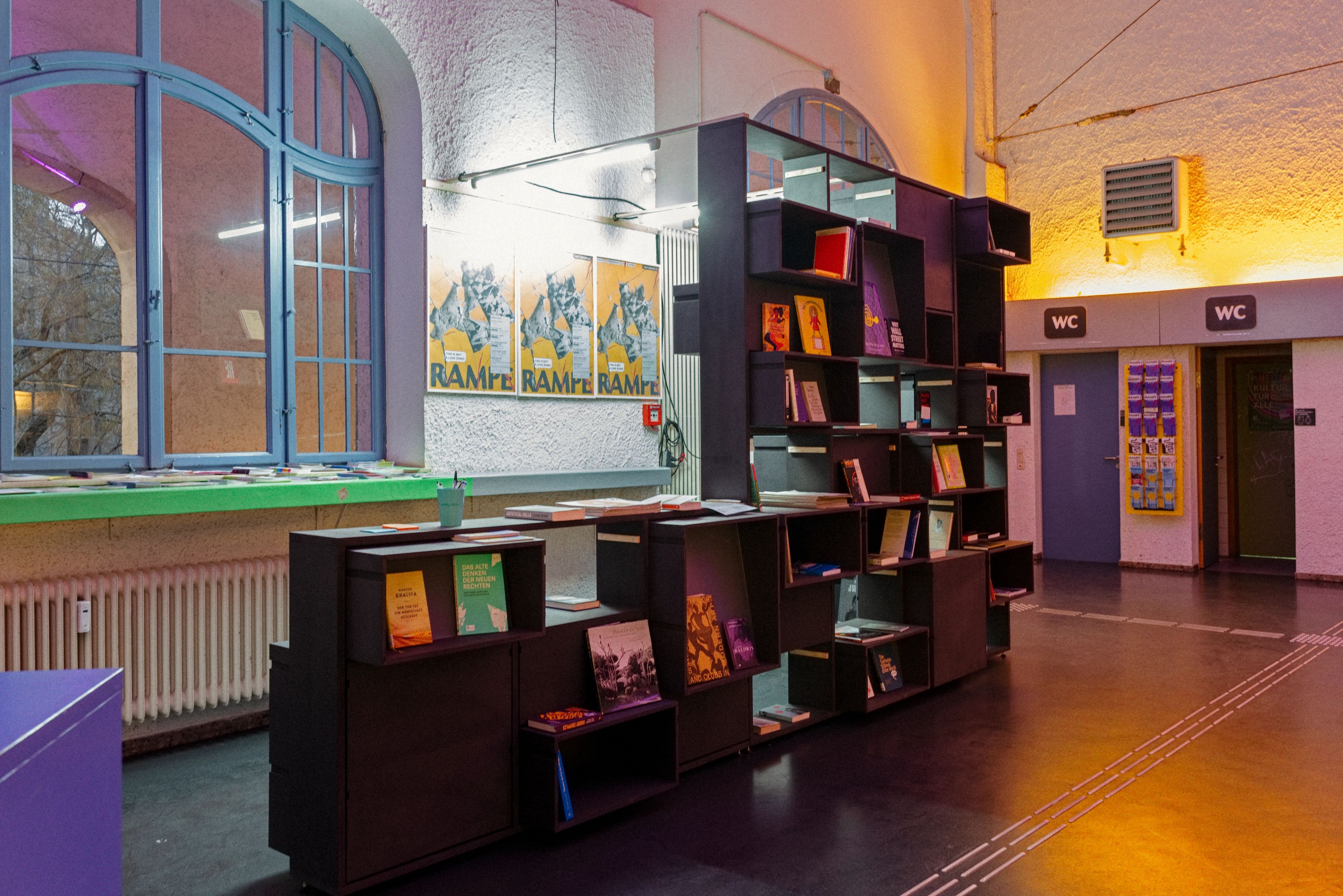A black modular bookshelf filled with books stands in a public interior with a large arched window and WC signs.