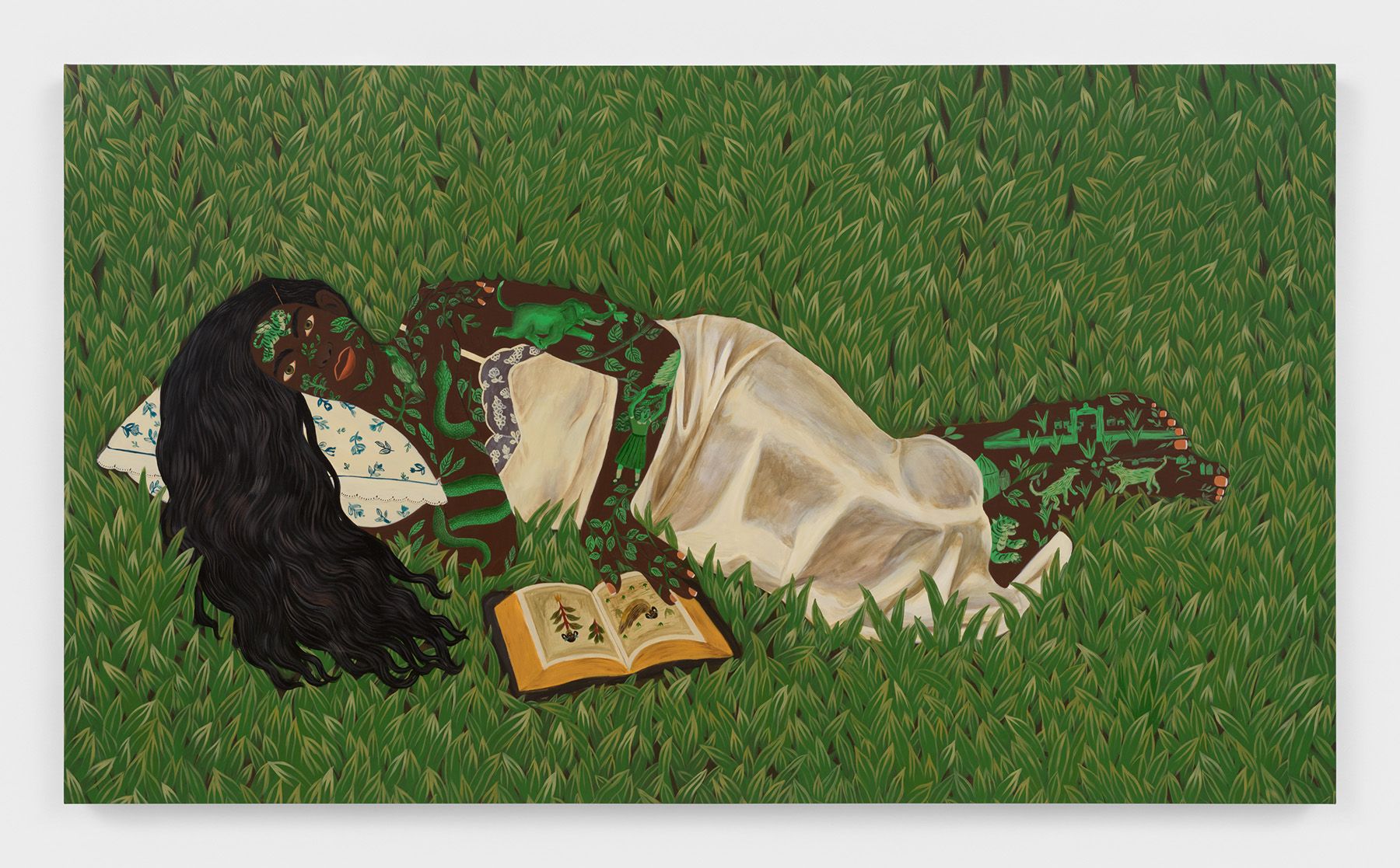 A painting of a person with long dark hair and dark skin patterned with green leaves and creatures, lying on a pillow in green grass with an open book.