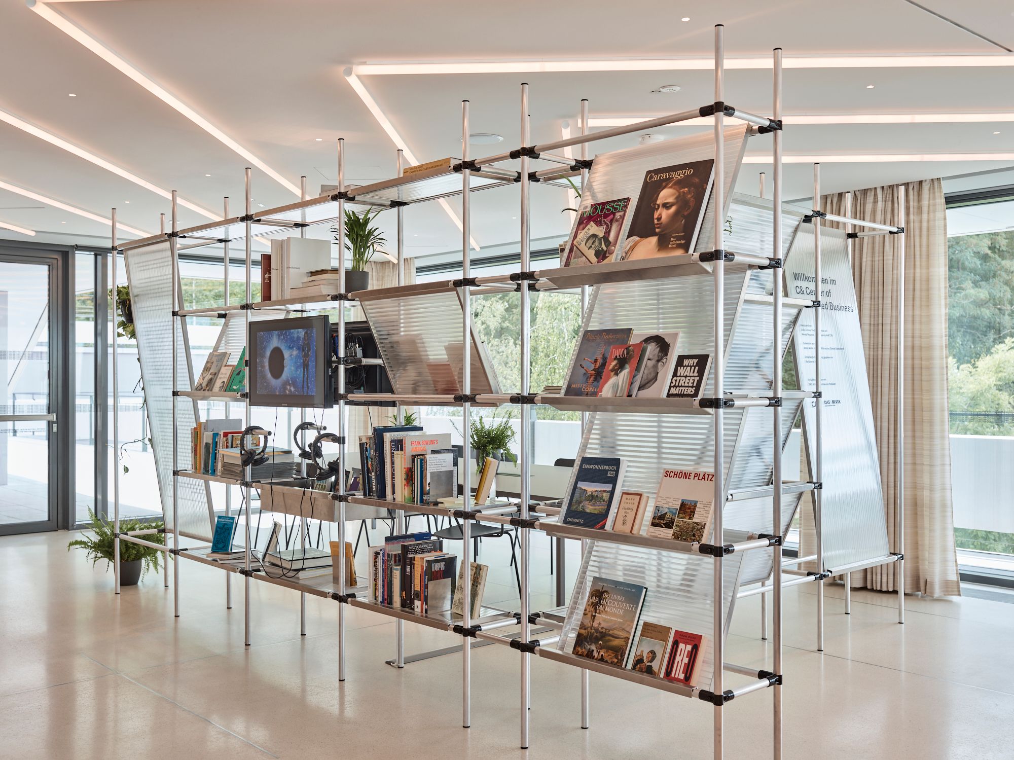 A modern, multi-tiered metal shelving unit acts as a room divider, displaying books, magazines, plants, and a monitor in a bright room.