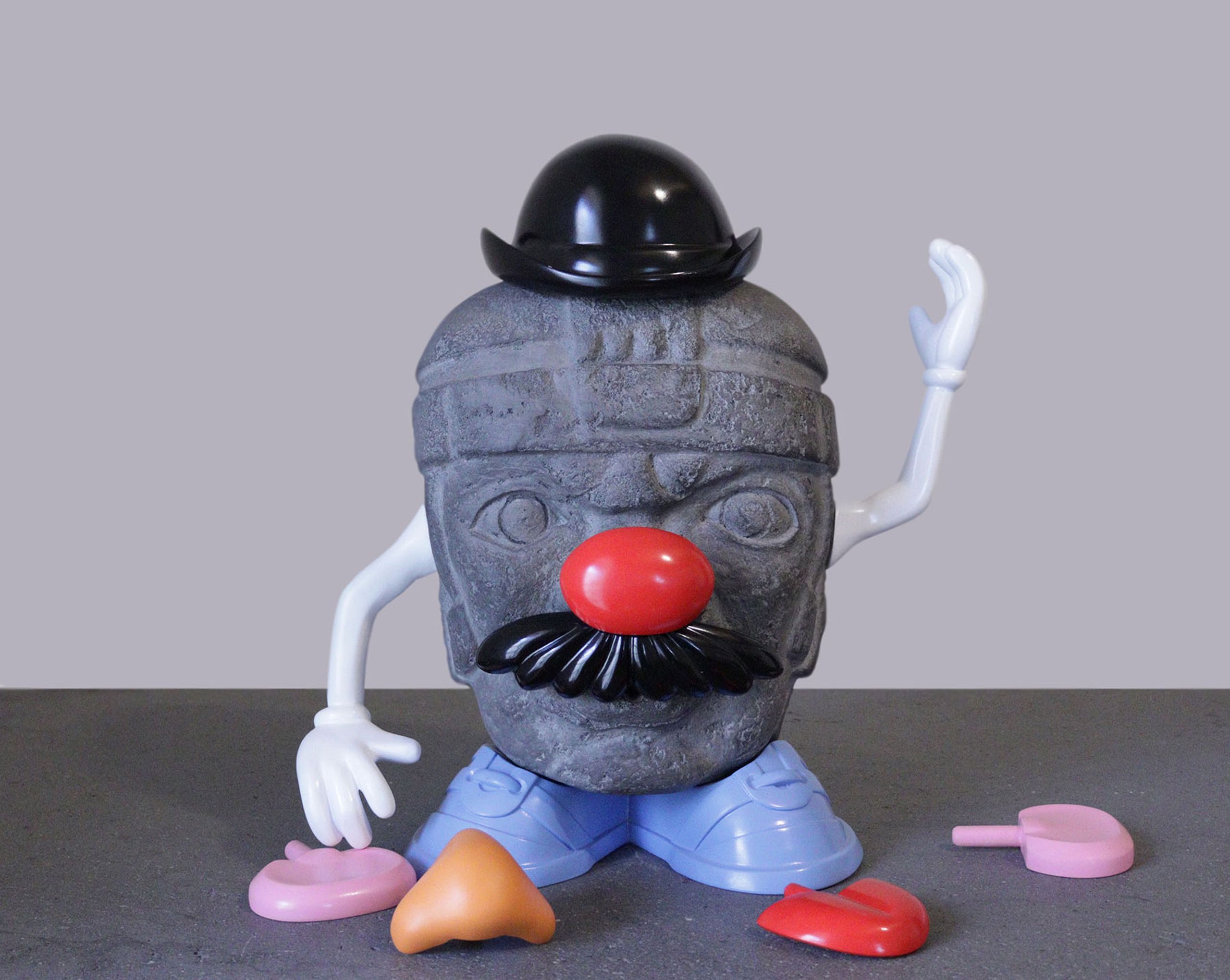An Olmec head sculpture fitted with Mr. Potato Head accessories, including a bowler hat, red clown nose, black mustache, white arms, and blue feet, with other plastic pieces on the ground.