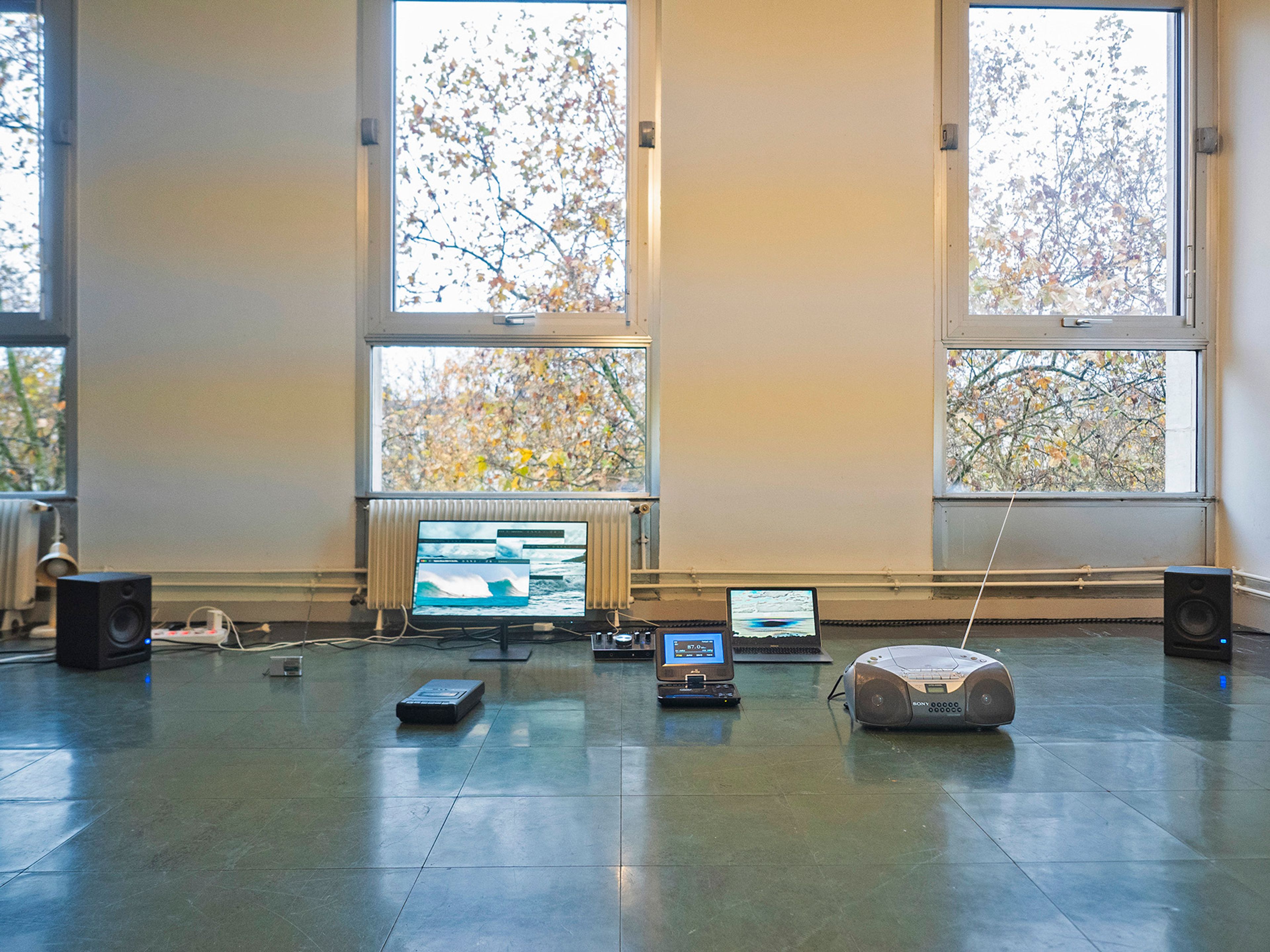 An array of electronic devices, including a monitor displaying glitch art, laptops, speakers, and a boombox, laid out on a dark floor in a room with large windows.