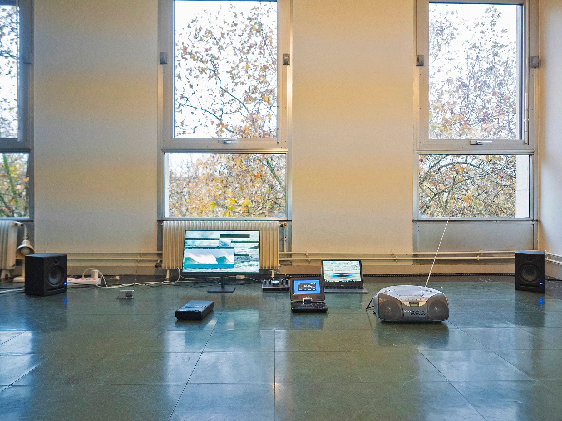 An array of electronic devices, including a monitor displaying glitch art, laptops, speakers, and a boombox, laid out on a dark floor in a room with large windows.